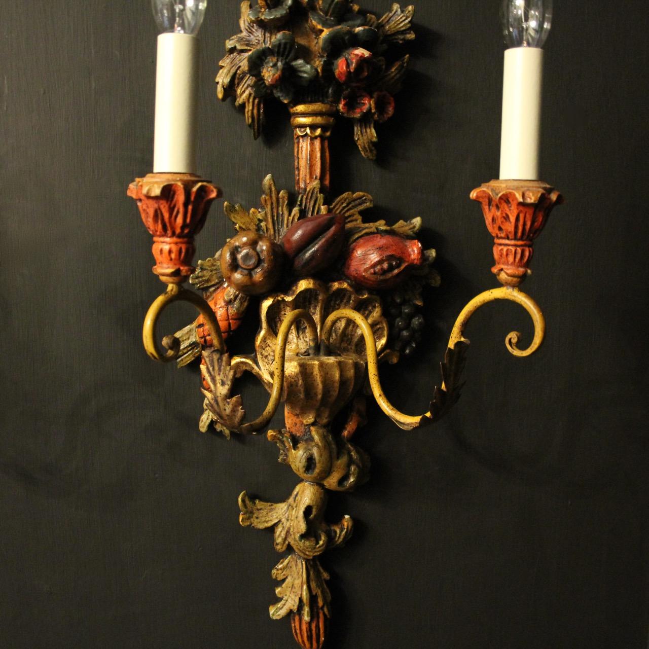 A decorative Italian polychrome and toleware painted carved wood twin arm wall lights, the metal leaf scrolling arms with carved wooden candle sconces, issuing from a ornately carved decorative floral and fruit clad carved wooden polychrome