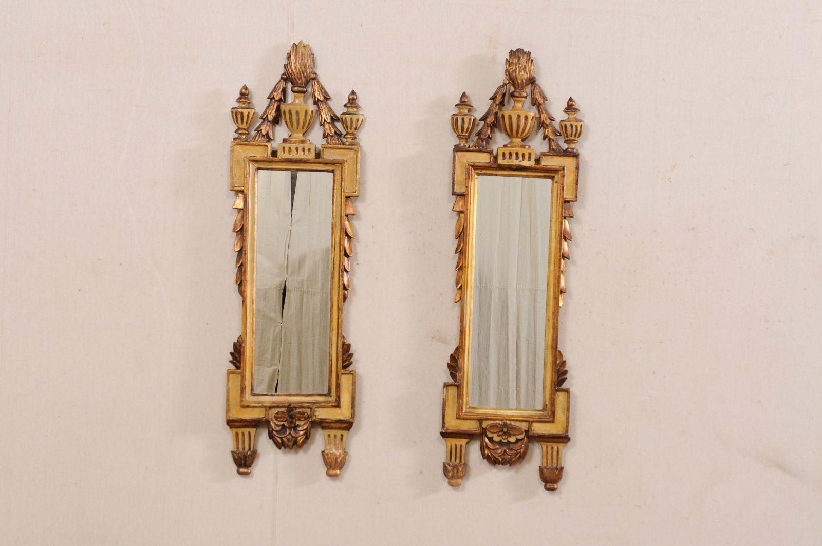 An Italian pair of Neoclassical wall mirrors from the 19th century. These antique mirrors from Italy feature Pots à Feu (Fire Urn) at their crest peaks with swagged garland draping downward to their shoulders adorn with raised finials. The mirrors