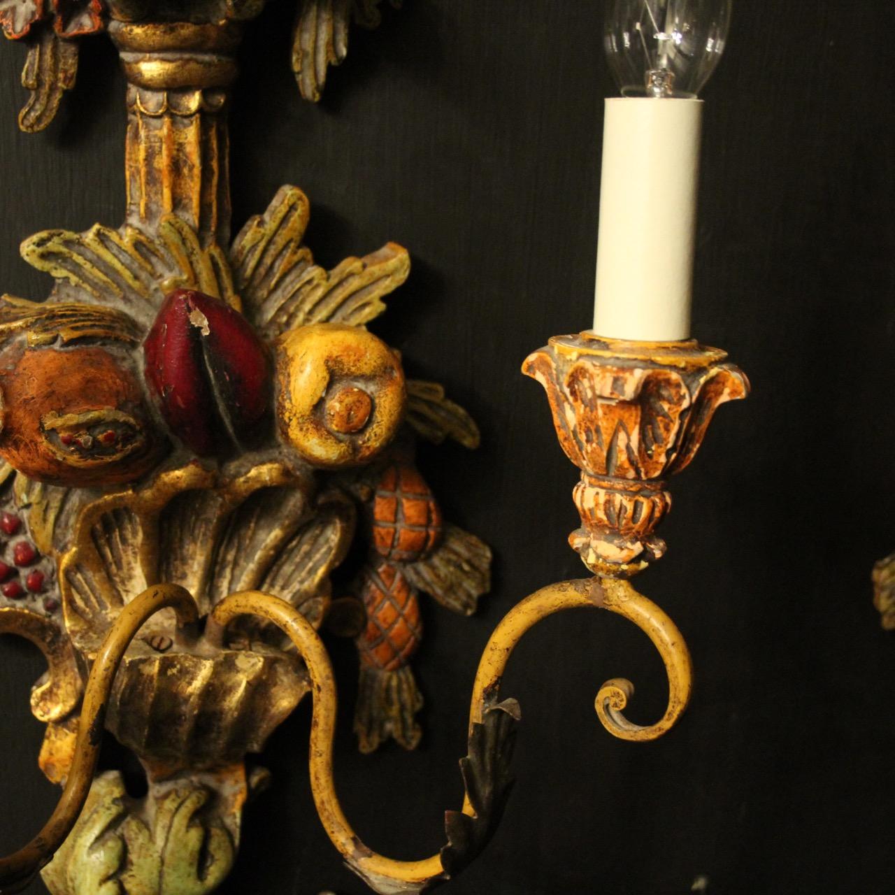 A decorative Italian polychrome and toleware painted carved wood twin arm wall lights, the metal leaf scrolling arms with carved wooden candle sconces, issuing from a ornately carved decorative floral and fruit clad carved opposing wooden polychrome