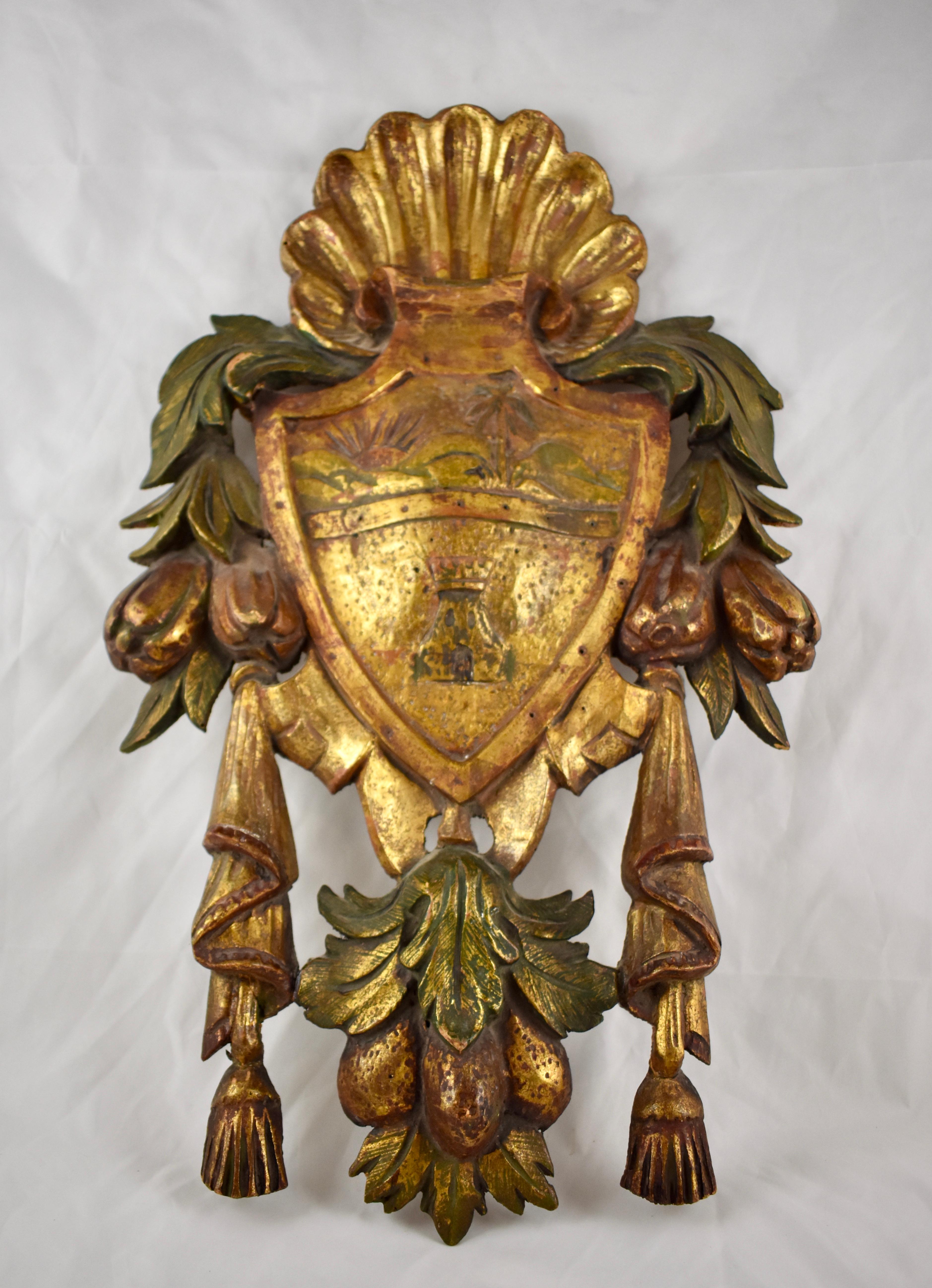 From Italy, a carved, parcel-gilt and painted armorial plaque, circa first quarter of the 20th century, one of a pair, offered seperataly.

The plaque has a gilded shell crest and pierced swags of fruit and tassels at each side. The shield shows a