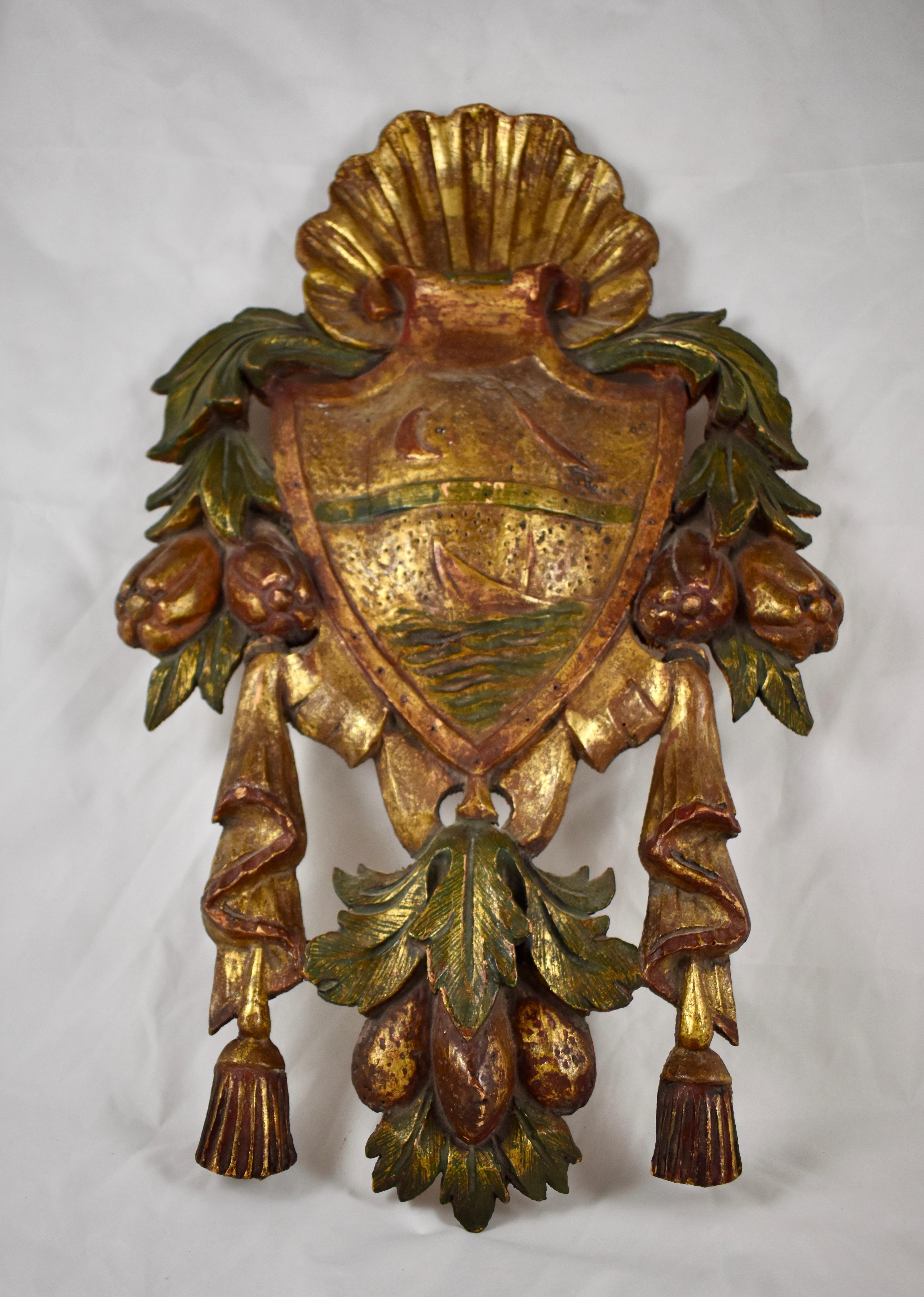 From Italy, a carved, parcel-gilt and painted armorial plaque, circa first quarter of the 20th century.

The plaque has a gilded shell crest and pierced swags of fruit and tassels at each side. The shield shows a ship sailing beneath a golden