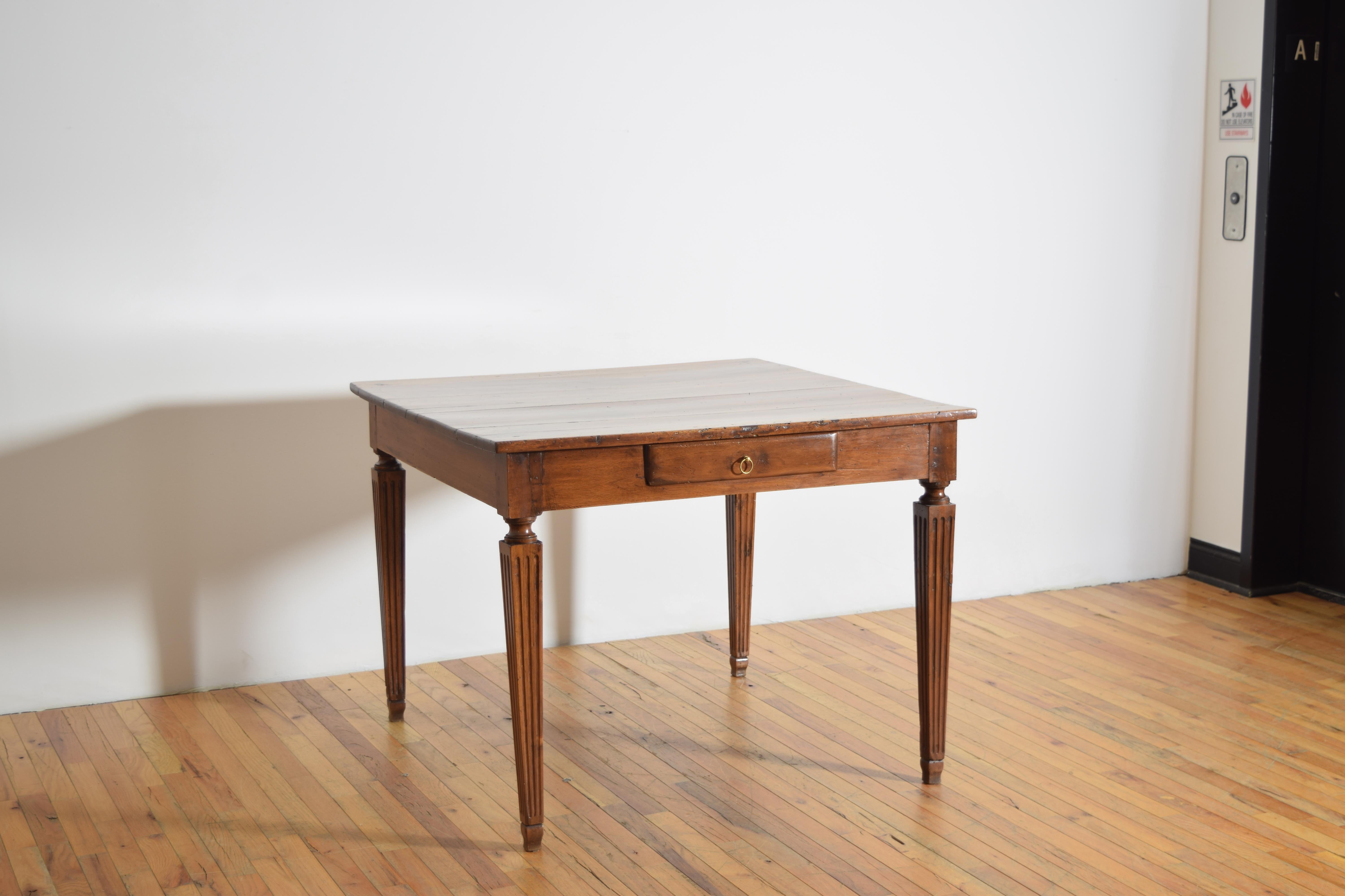 a center, kitchen, or games table, having a square top and shallow apron housing one deep drawer with a brass ring pull, the top raised on square tapering and fluted legs atop small shaped feet