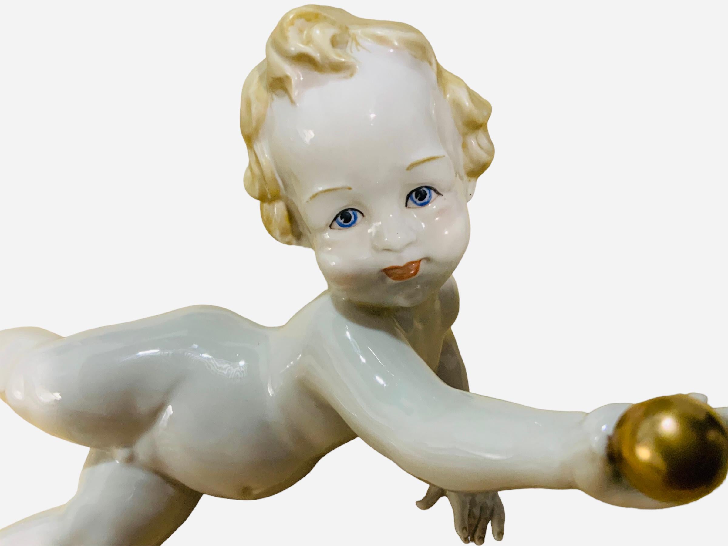 This is an Italian Capodimonte Porcelain Baby Boy Piano Figurine. It depicts a joyful nude baby boy with blonde curly hair and large blue eyes laying down in one side and holding a gold ball with one hand. Below the baby base is hallmarked G with a