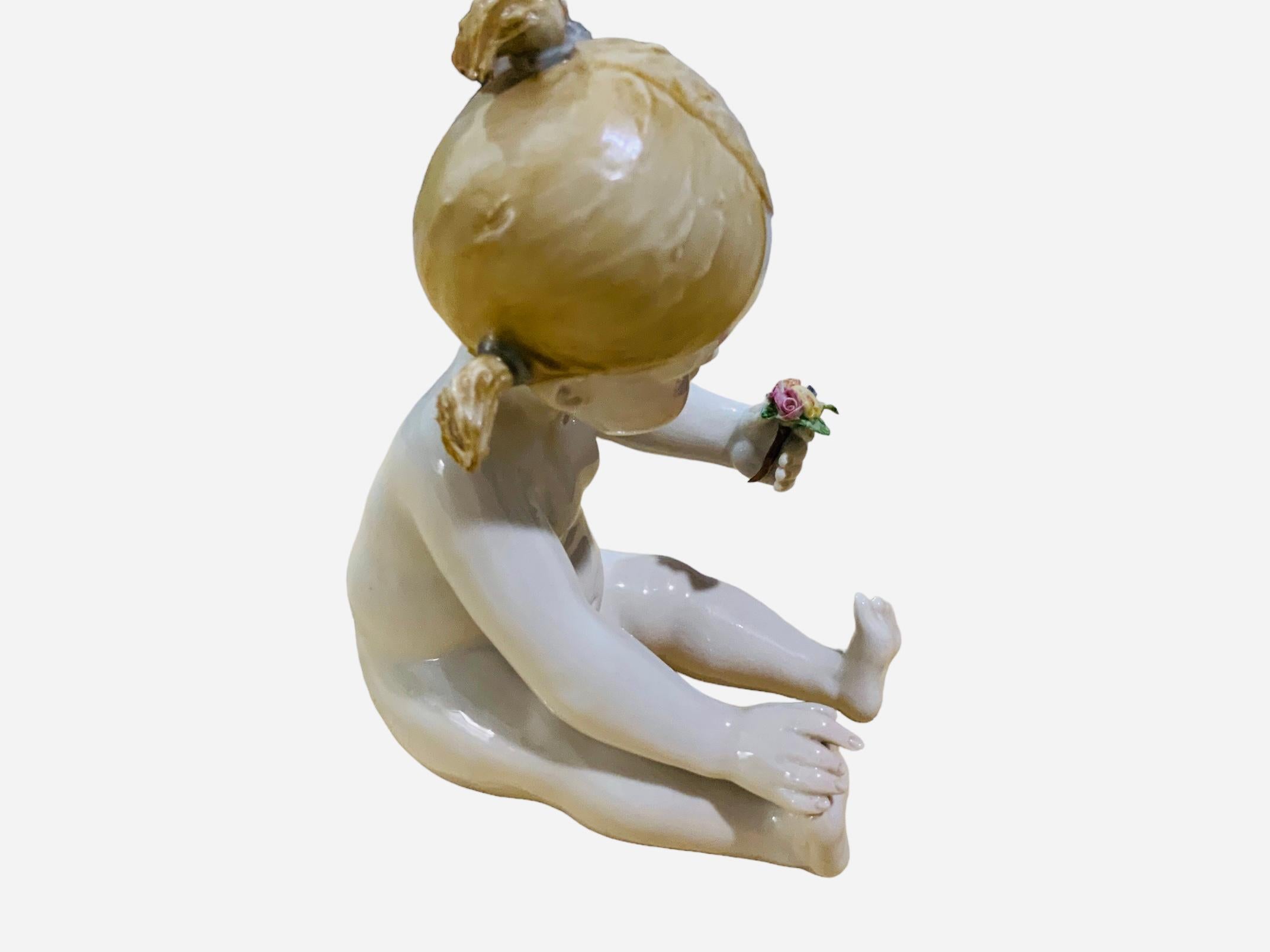 This is an Italian Capodimonte Porcelain Baby Girl Piano Figurine. It depicts a nude baby girl with large blue eyes and blonde hair arranged with ponytails sitting down admiring a bouquet of flowers that she is holding with one of her hands. Below