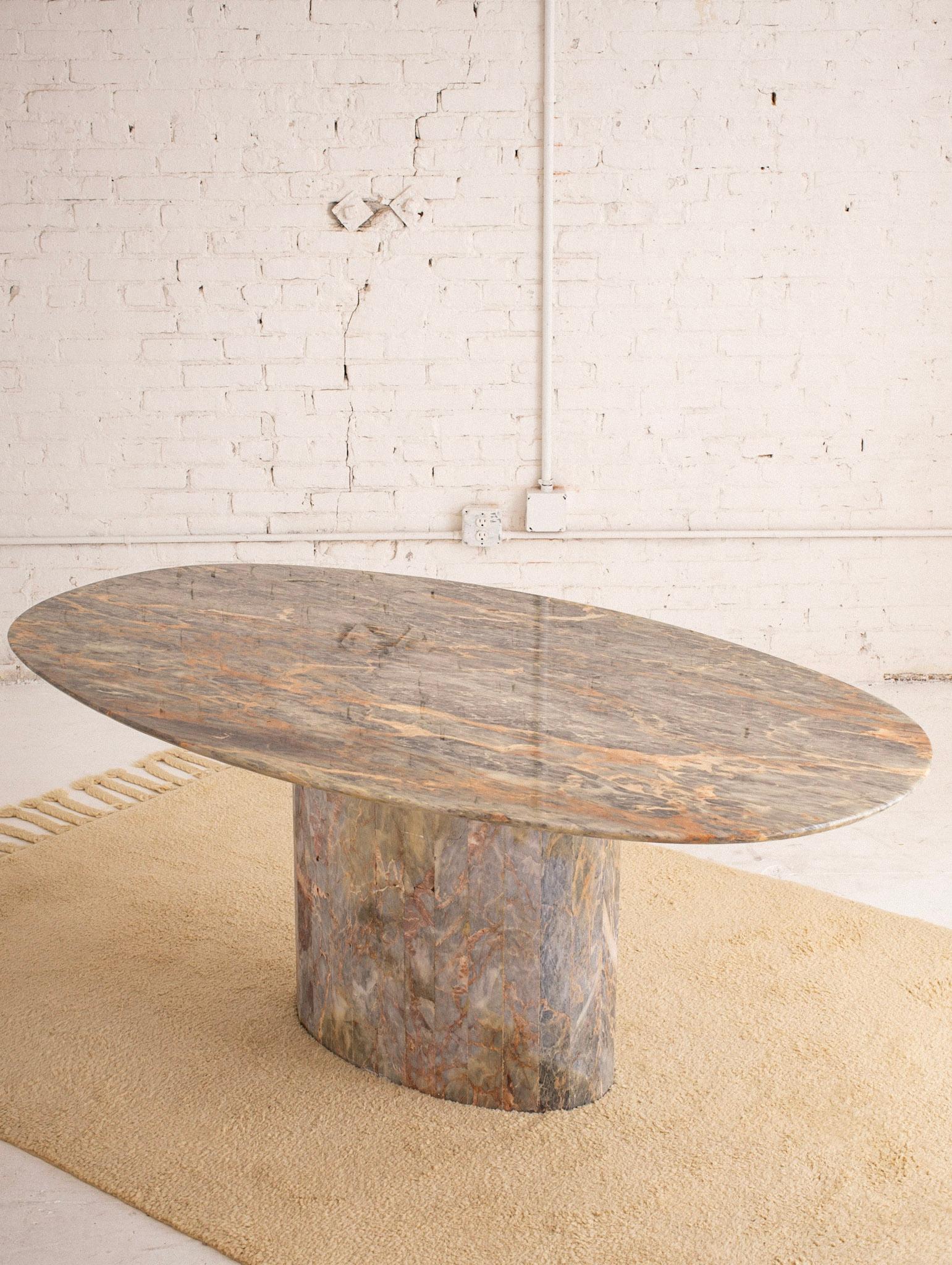 An Italian marble dining table. Oval table top rests on a cylindrical marble base. Vivid color variation throughout ranging from greens and greys to beiges and pinks.