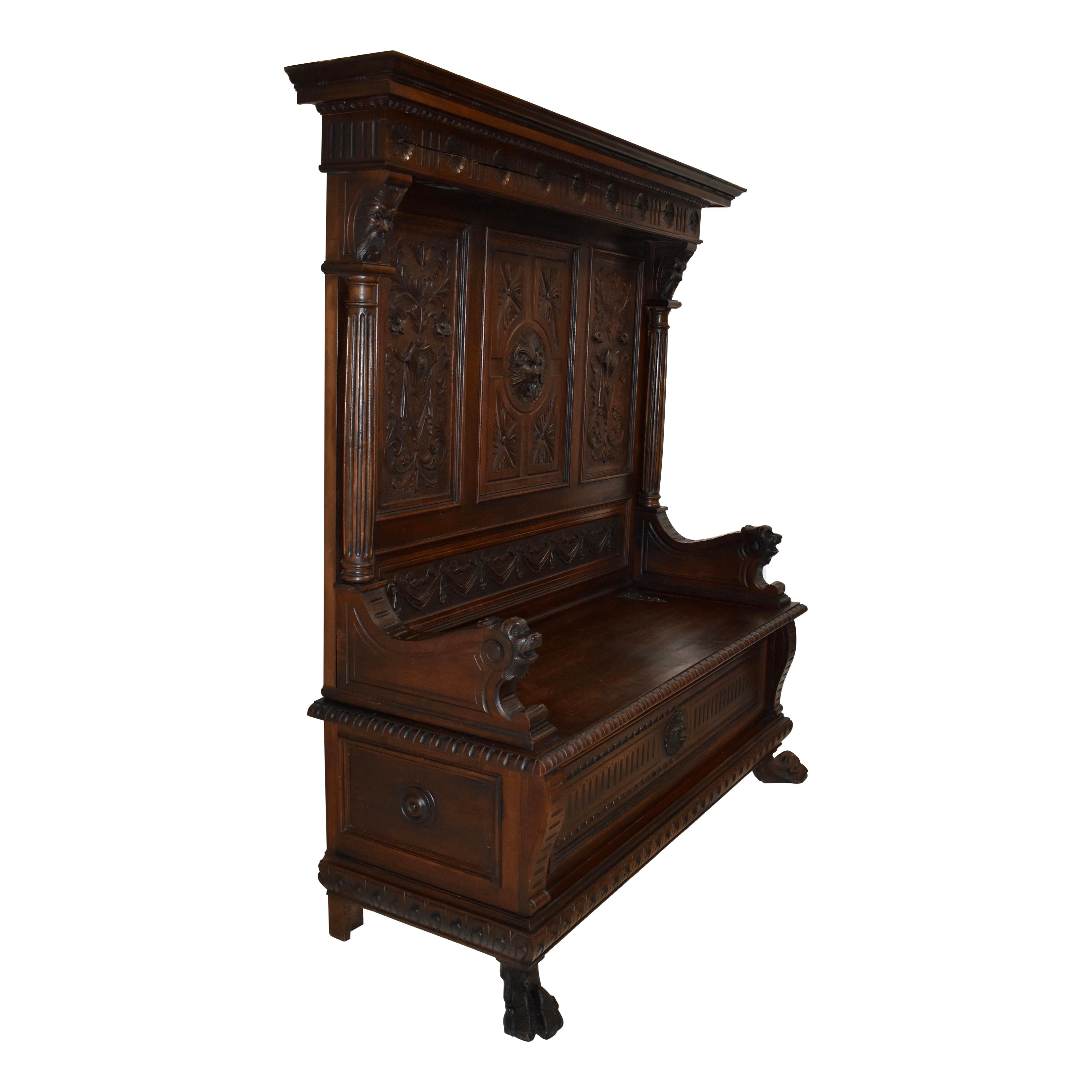 With elaborate carvings of the Renaissance Revival style, this walnut bench features a molded cornice over a frieze with floral roundels, a high back, scrolled arms, upright fluted columns, a bench seat on iron strap hinges that opens to divided