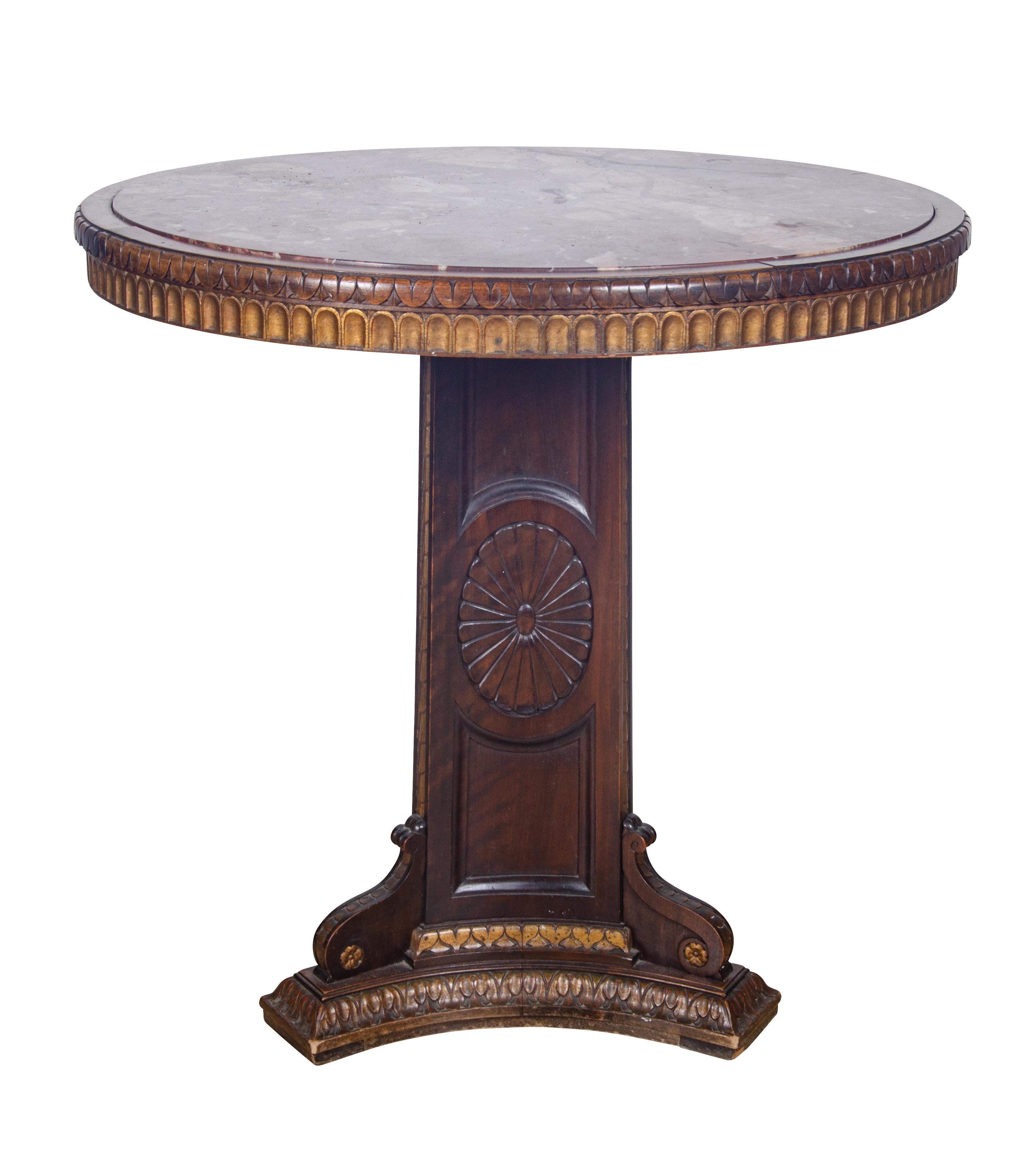 With an inset circular marble top with a rich red color set in a conforming wood border with carved details , the base carved and with subtle gilded details and triangular shaped base.