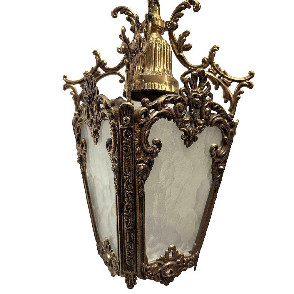 Este exquisito farolillo colgante de estilo rococó italiano es una auténtica obra de arte, fabricada en latón macizo. Irradia una cálida pátina que resalta su rica historia y elegancia. La lámpara presenta florituras rococó, como intrincadas