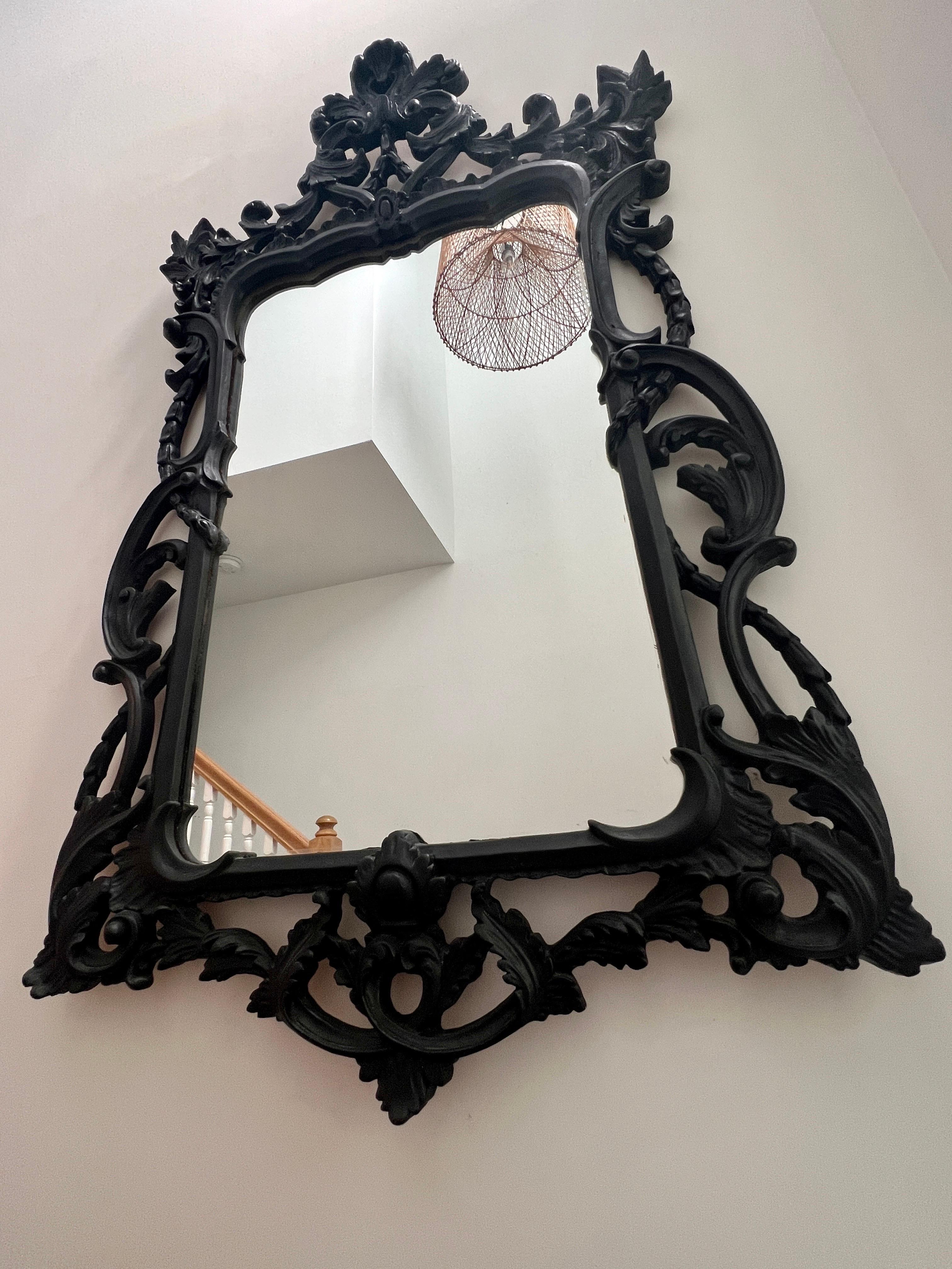 Large Hollywood Regency mirror with traditional Rococo style carved wood frame. Mirror has shield design featuring a series of hand carved scrolls with foliage details. The vintage black finish has a slightly distressed quality which gives the
