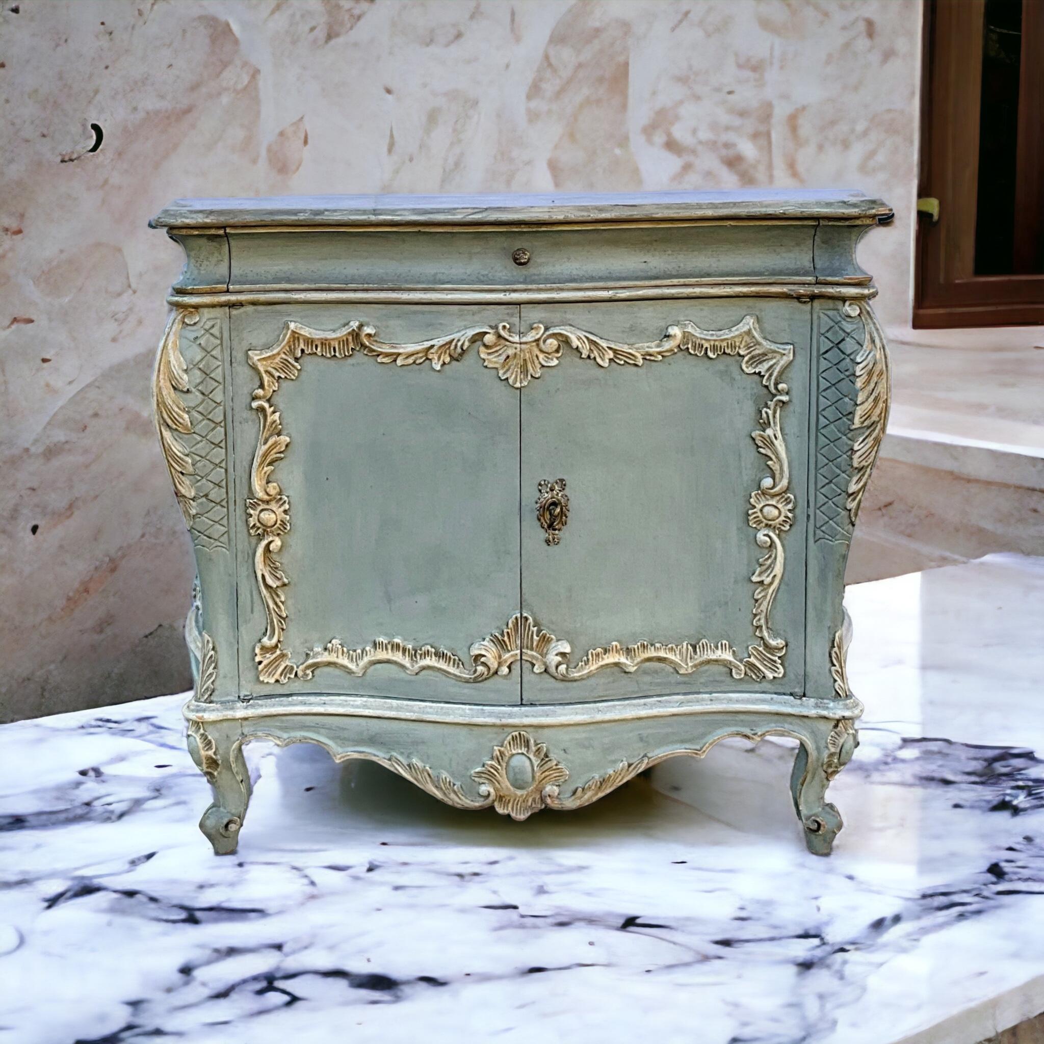 This is an Italian Rococo style heavily carved commode in a French blue with ivory embellishments. The top is a hand painted faux marble. It is marked, “Made in Italy.” It is a mid-century piece with a single drawer over two doors.