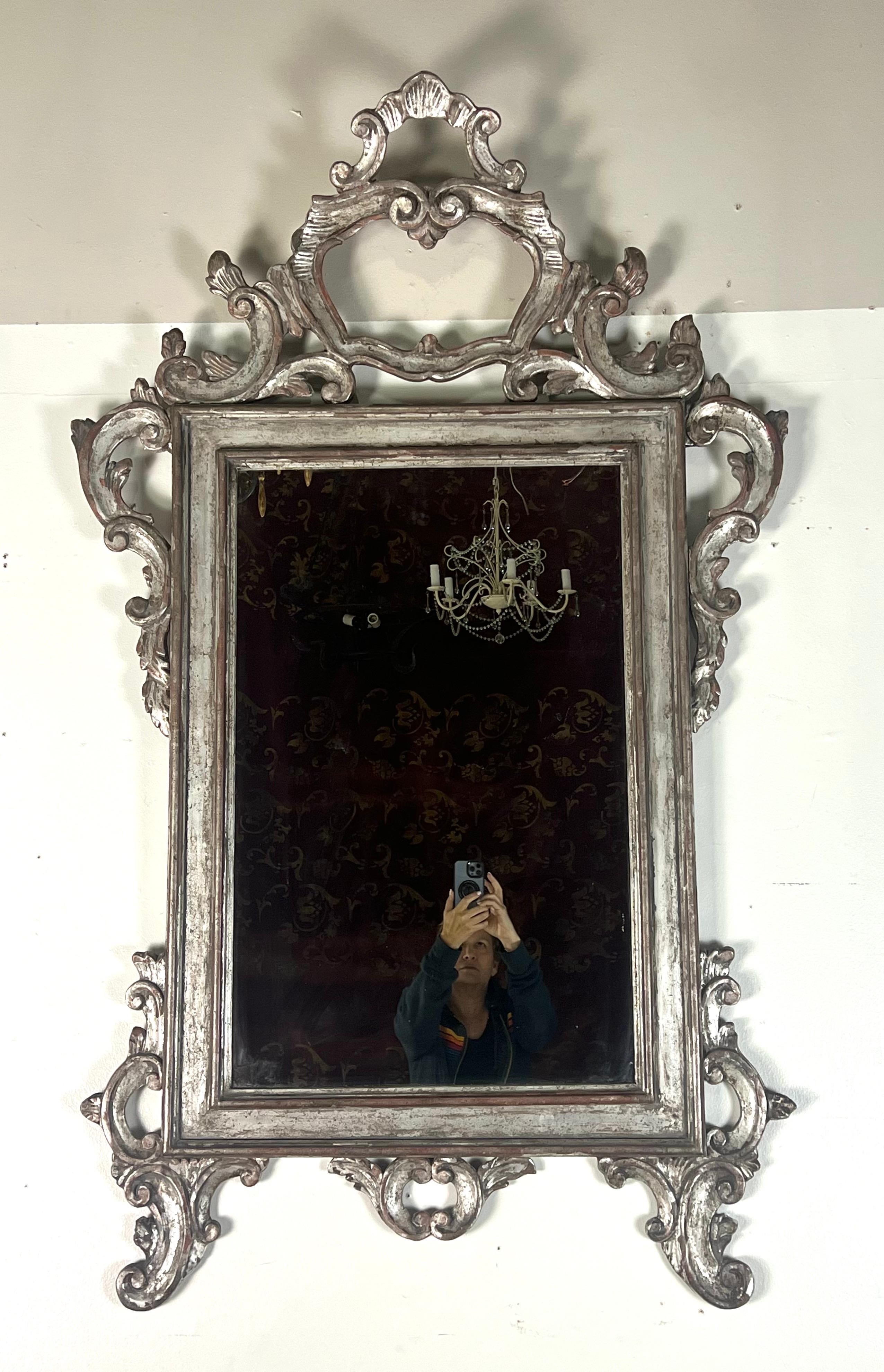 Dies ist ein kunstvoller Spiegel im Rokoko-Stil mit einem aufwendig geschnitzten Rahmen.  Der Rahmen zeichnet sich durch komplizierte Schnörkel und dekorative Schnörkel aus, vor allem oben, wo ein großartiges, wappenartiges Design Höhe und Eleganz