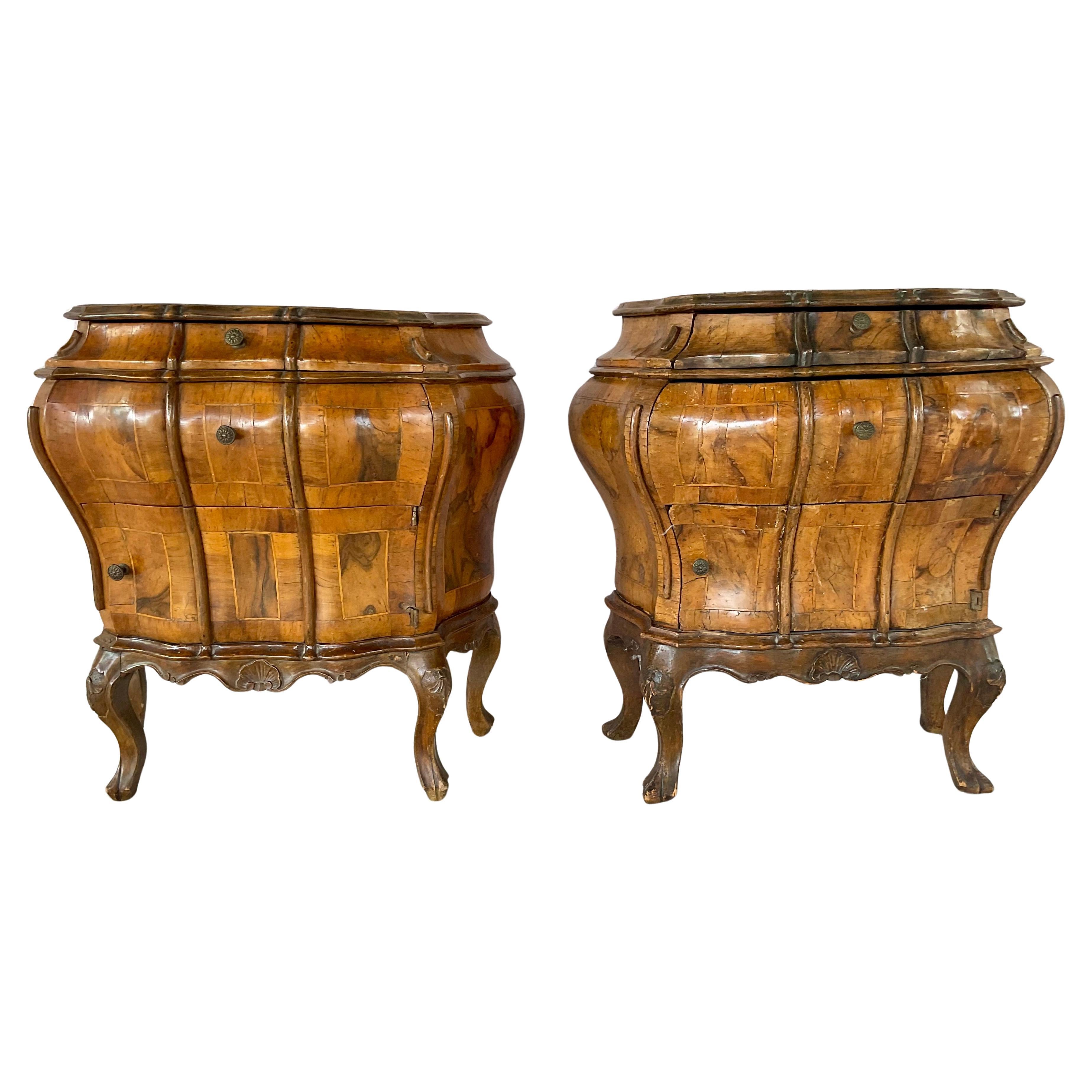 A Pair of Italian Rococo Style Bombe Commodes crafted from richly grained burl walnut, each with a curvaceous serpentine form and three drawers. Commodes feature delicately carved aprons and subtle shell motifs and rest on cabriole legs. The patina
