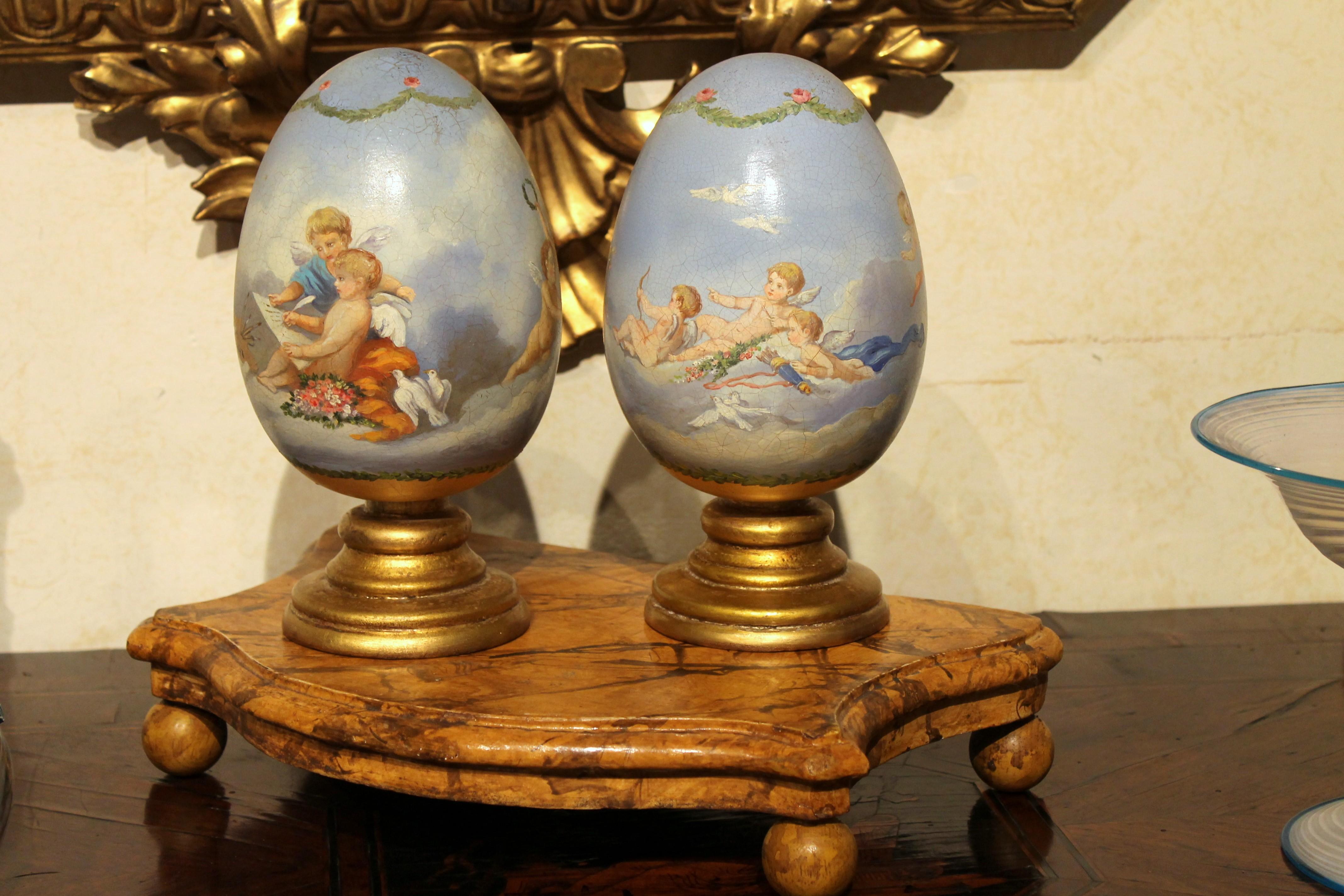 This pair of Italian romantic terracotta eggs painted in the round on giltwood stands are a lovely decorative object, hand painted with extreme attention to details features putti and cherubs lying on white fluffy clouds in a blue sky. 
A putto is