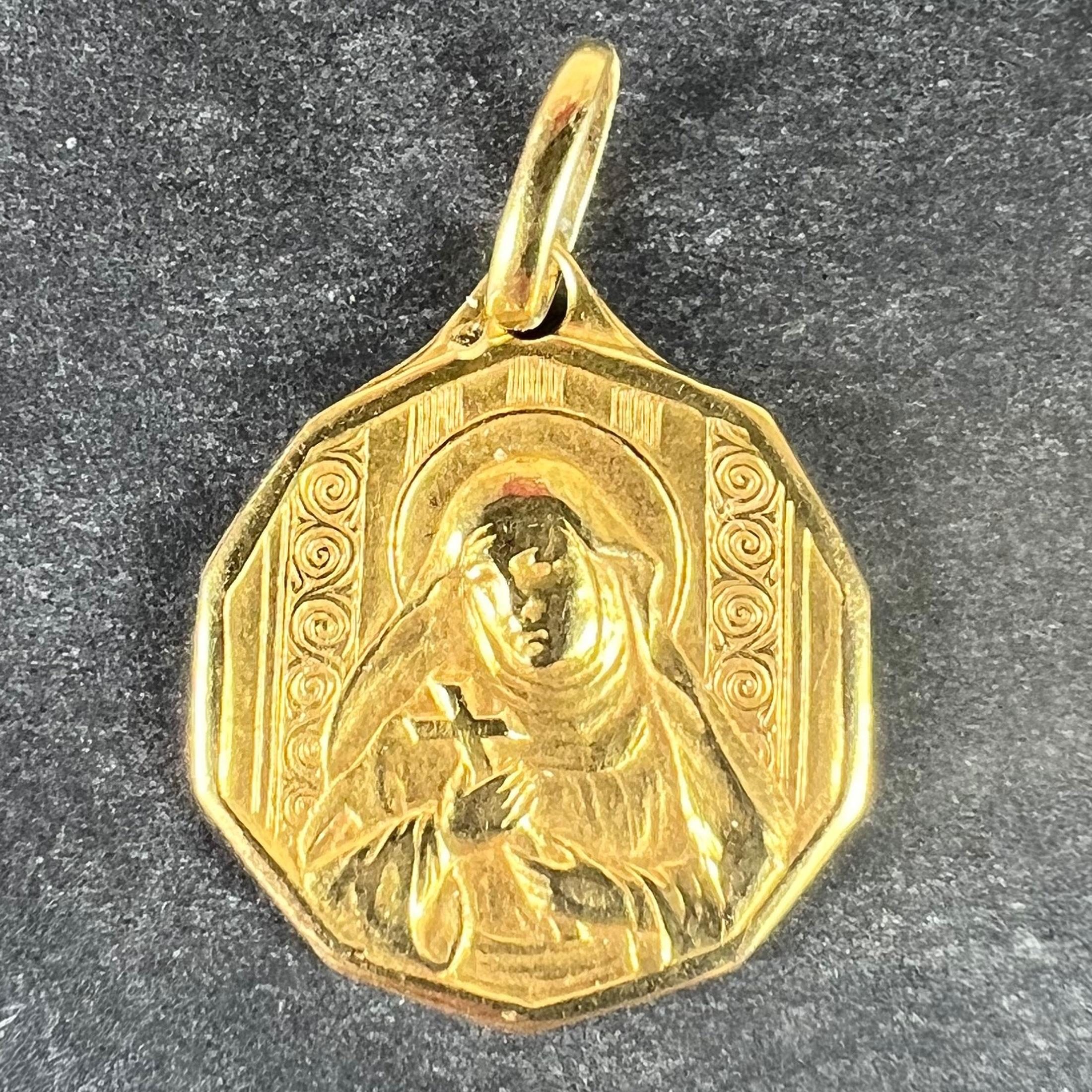 An Italian 18 karat (18K) yellow gold charm pendant designed as a medal depicting Saint Therese with a halo holding a crucifix with a background of stripes and swirls to represent roses. Stamped with the owl mark for 18 karat gold and French import