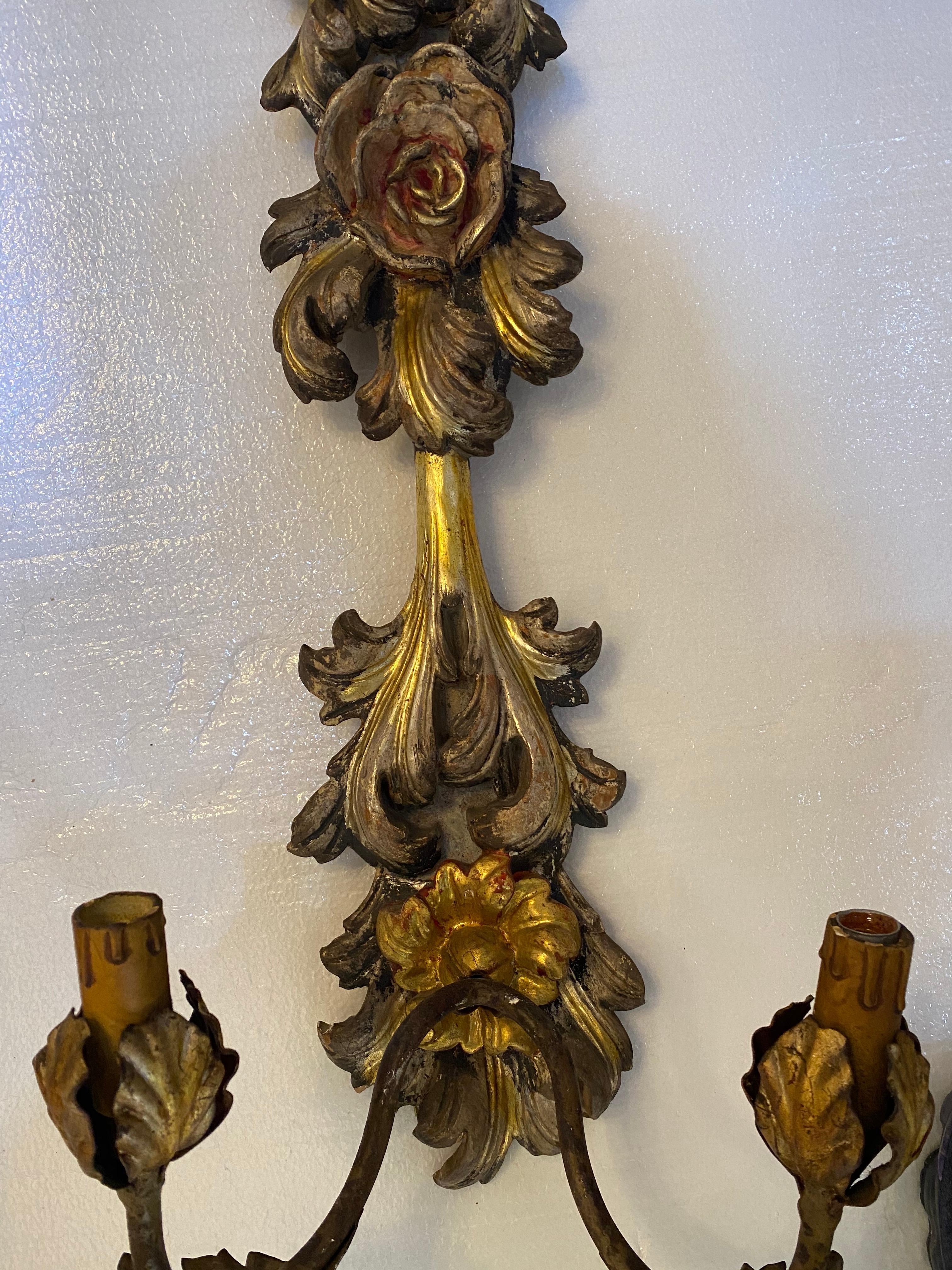 Two lights ea. Italian two-light wood sconces, early 20th c., parcel gilt and polychrome painted wall mounts, having relief carved foliate and rose motif, supporting two patinated metal arms ending in petal bobeche and faux candles.