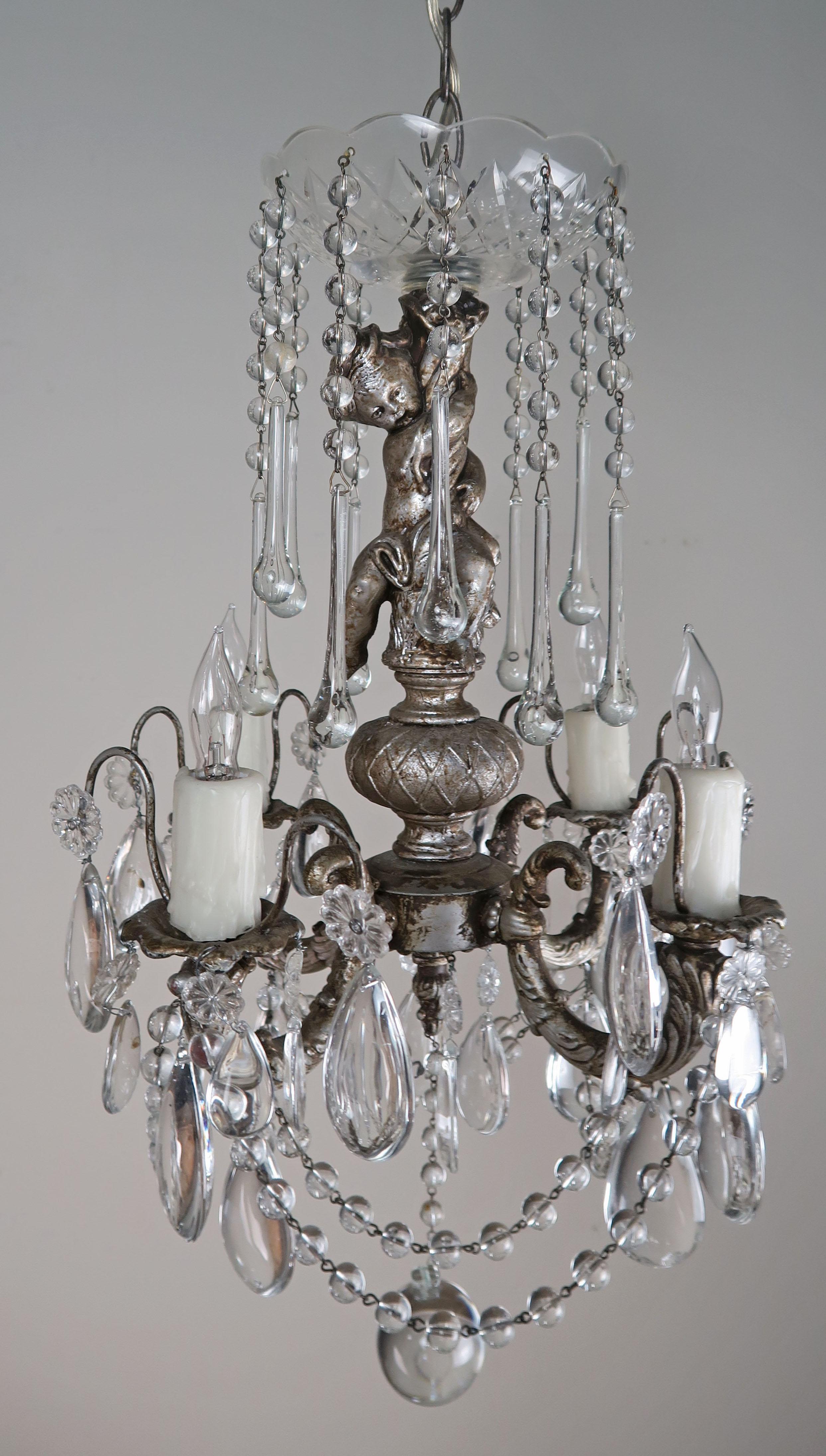 Four-light silvered gilt metal chandelier adorned with crystals and crystal garlands throughout. A charming sweet faced cherub is depicted as part of the column of the chandelier. The fixture is newly rewired with drip wax candle covers. Chain and