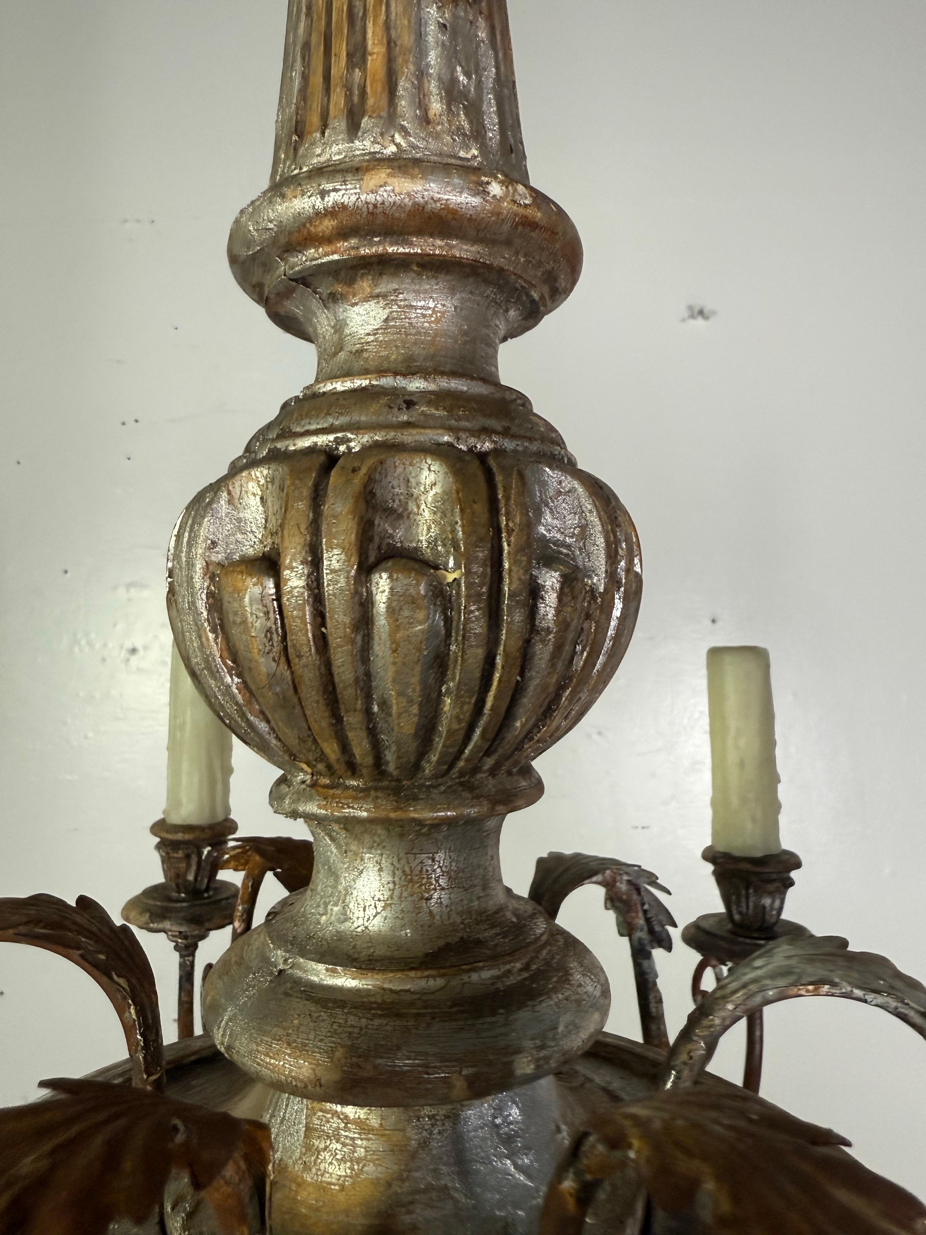 Un lampadario italiano splendidamente invecchiato che presenta una colonna centrale in legno argentato intagliato con dettagli scanalati e cangianti. Sei bracci di ferro forgiati a mano si estendono in graziose volute, ognuna delle quali sostiene un