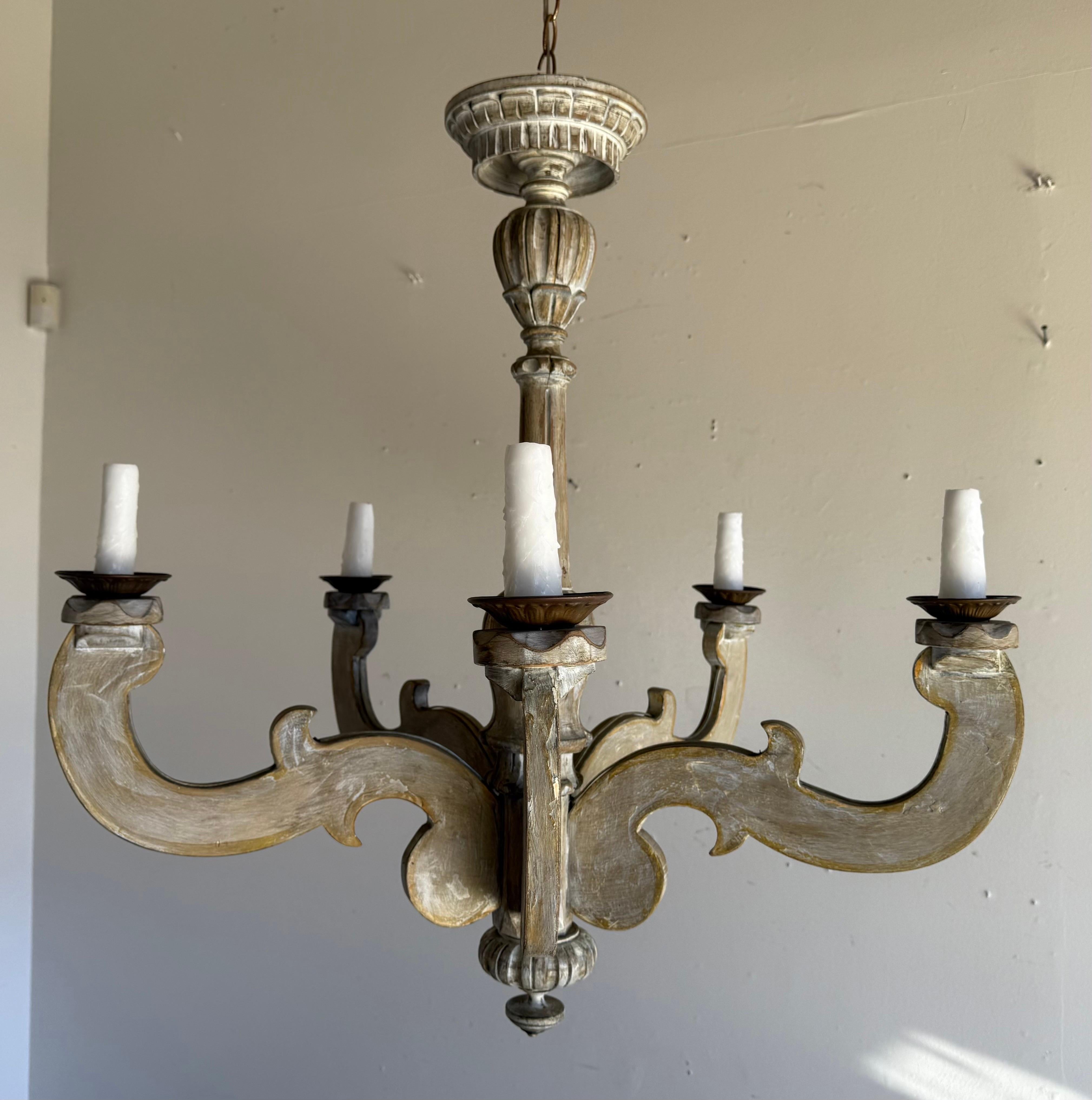 An elegant Italian six-light chandelier, mid-20th century, crafted in carved wood with gracefully scrolling arms and classical detailing. The frame is finished in a timeworn palette of soft white with hand-gilded accents, giving the piece a warm,