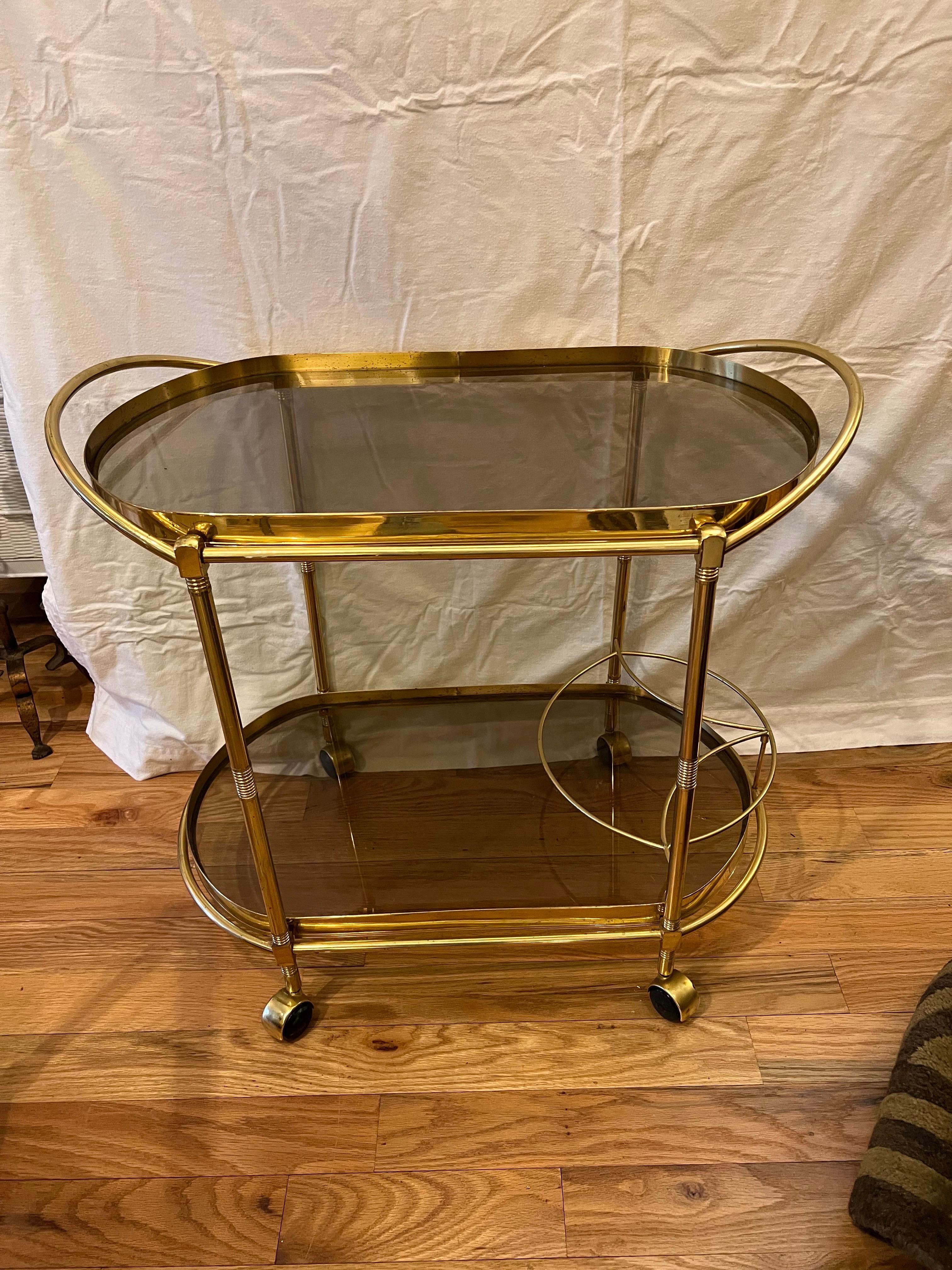Smoked glass two tiered oval bar cart. Most likely Italian with its sleek and sexy lines. This Hollywood Regency style smoked glass and brass two tier bar cart has a bottle holder on the lower tier. Elegant oval shape with a handle to push and brass