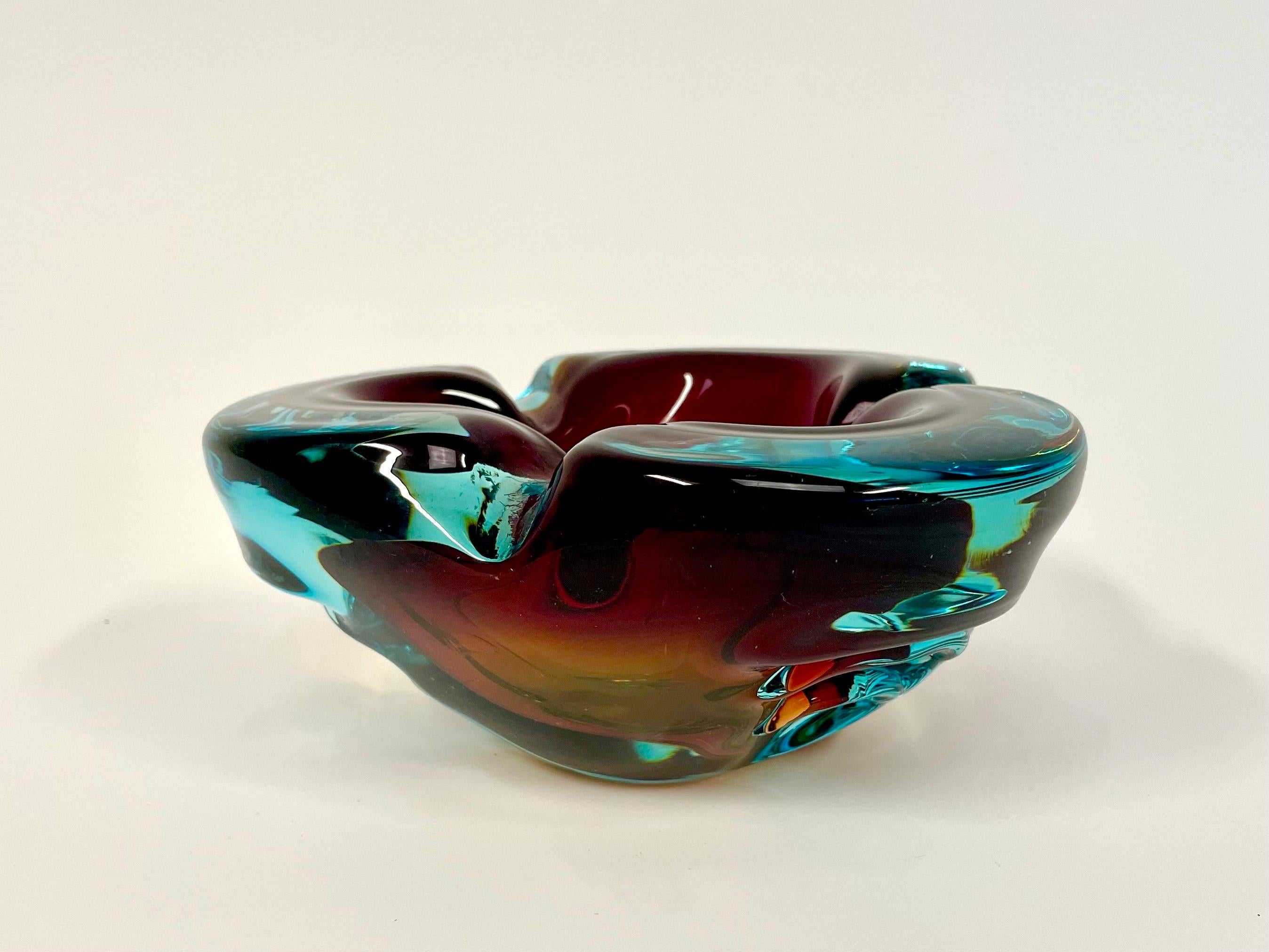 Il s'agit du lourd cendrier en verre d'art d'Alfredo Barbini, aux formes douces. Il est proposé dans une variante tricolore moisie en bleu méditerranéen avec des éléments bordeaux et un fond ambré. 

Il présente un design triangulaire avec des