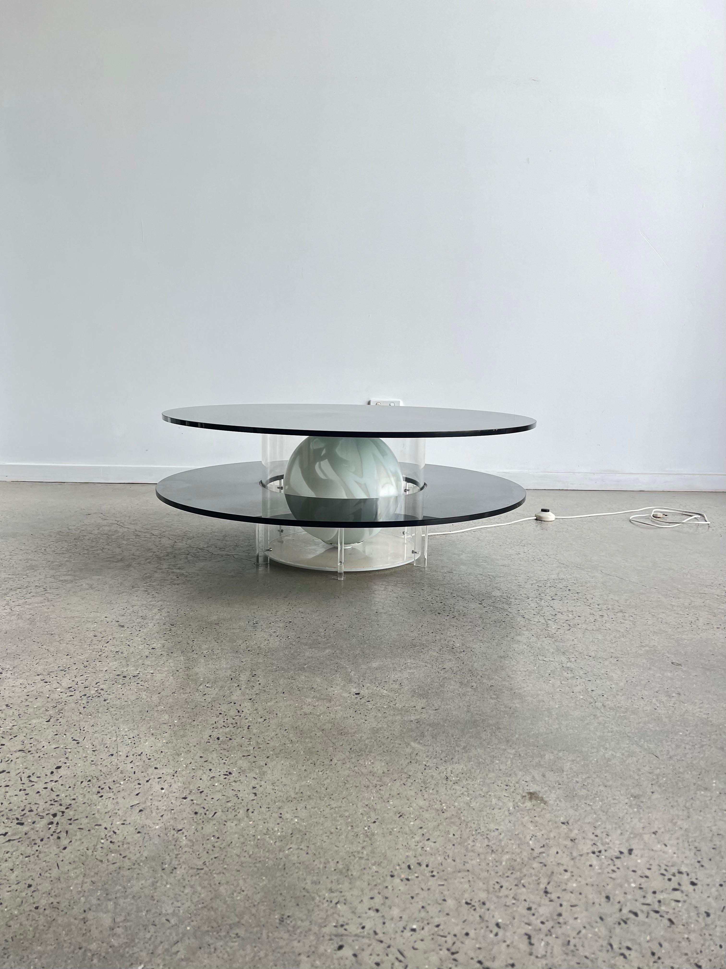 The Space Age Round Coffee Table with Smoked Glass, Plexiglass base, and inside Murano Glass Ball is a unique piece of furniture from the 1970s. The table features a round top made of smoked glass, which sits on a plexiglass base. The base has a