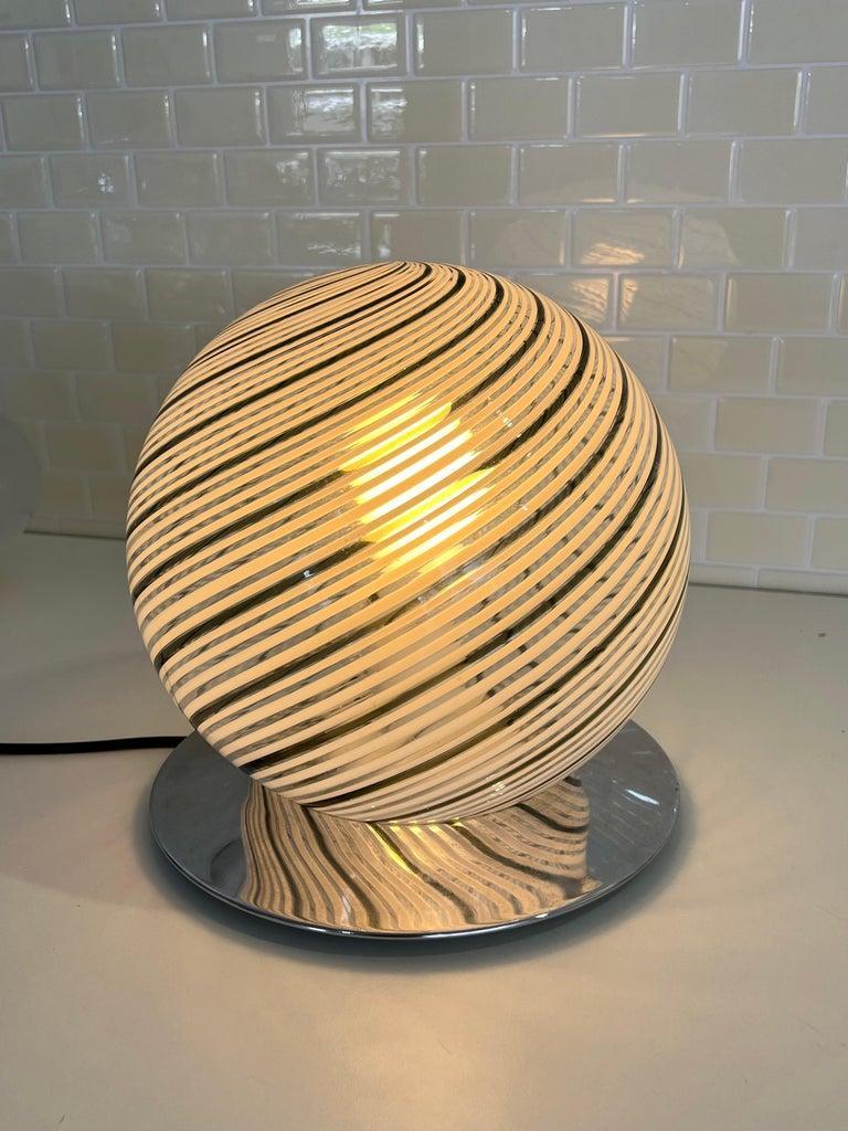 Die Lampe hat einen kugelförmigen Schirm aus Murano-Glas, der von erfahrenen Handwerkern in Italien handgefertigt wurde. Der Glasschirm hat eine schöne Textur und Farbe, die in jedem Raum ein warmes und einladendes Ambiente schafft.

Der verchromte
