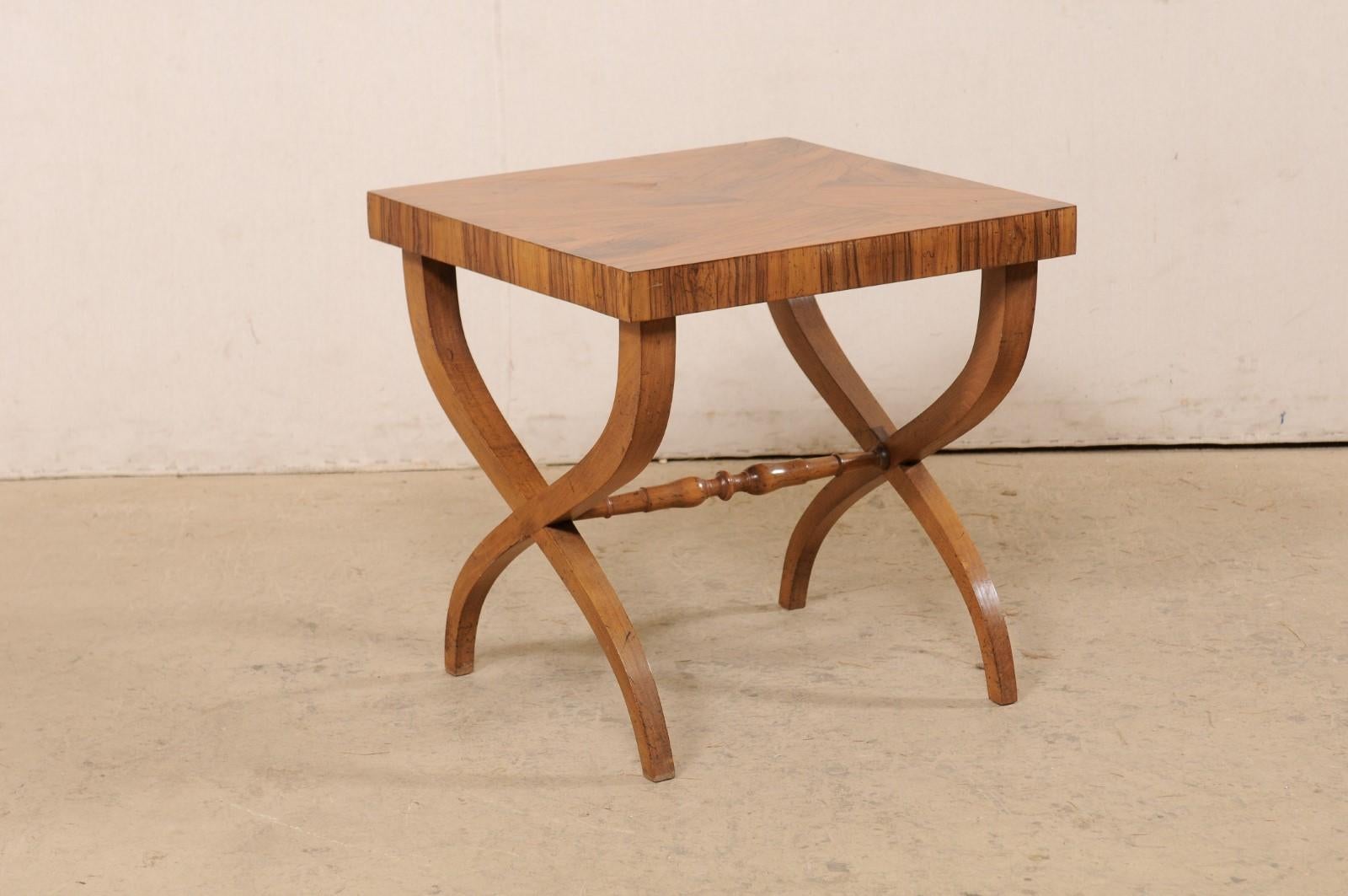 An Italian curule side table from the mid 20th century. This mid-century table from Italy has a square-shaped top of beautifully veneered olivewood, which is raised on a pair of curule (wavy x-shaped) wooden legs. The legs are braced at underside of