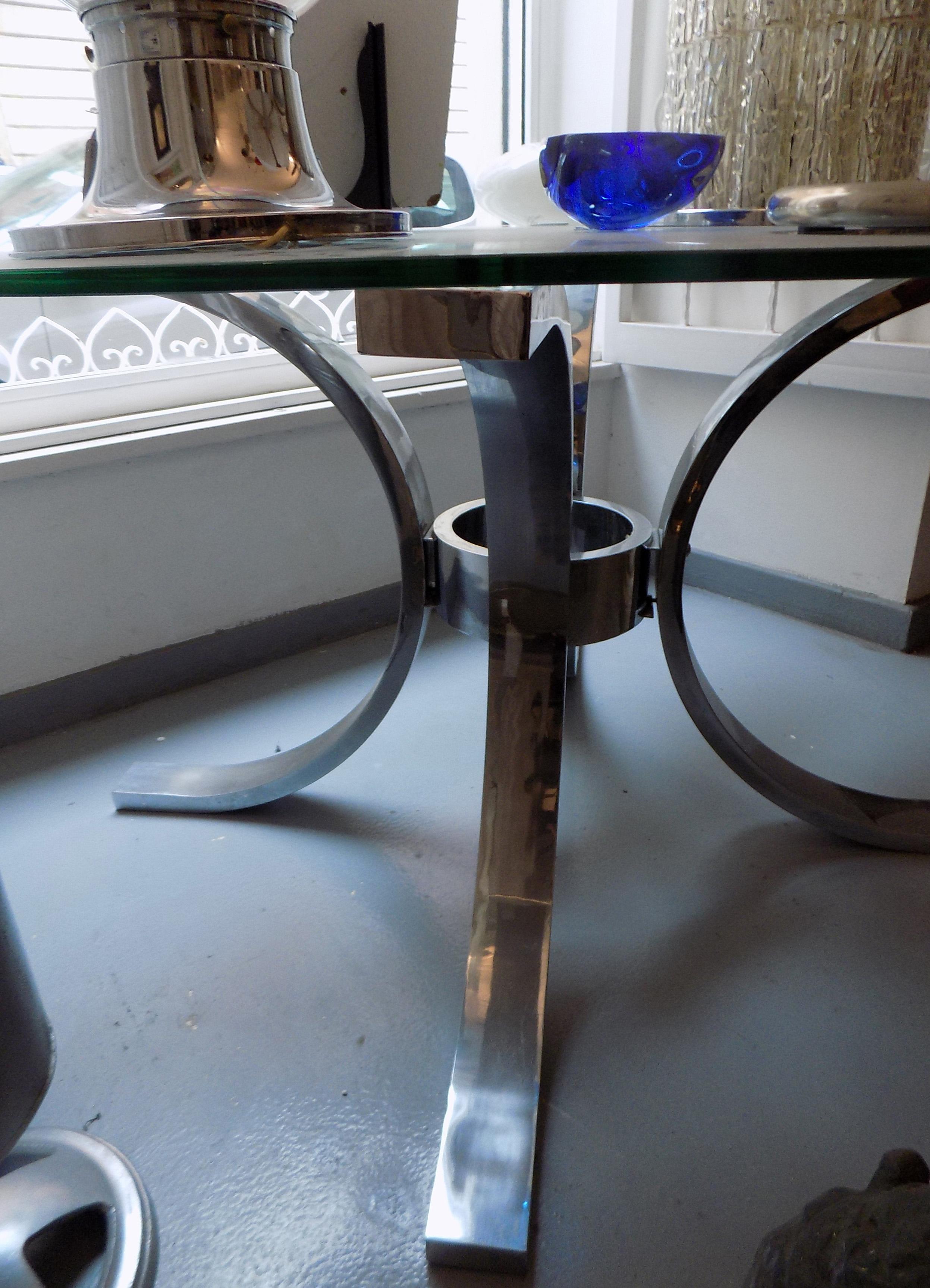 1970s table with glass top, and metal leg structure. The glass top is circular in shape, while the leg is made up of three arch-shaped elements, in the manner of Osvaldo Borsani.