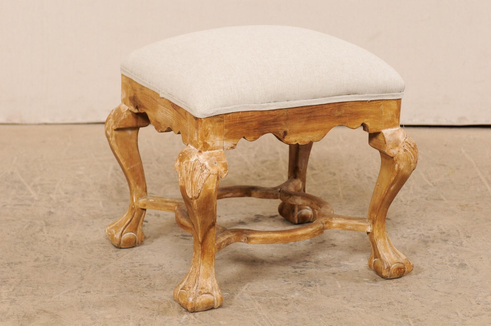 A vintage American carved-wood ottoman with upholstered seat. This stool, with its Italian inspired design, feature an upholstered seat above a valance carved skirt, and raised upon thickly carved legs, with profiled knees carved with shells, and