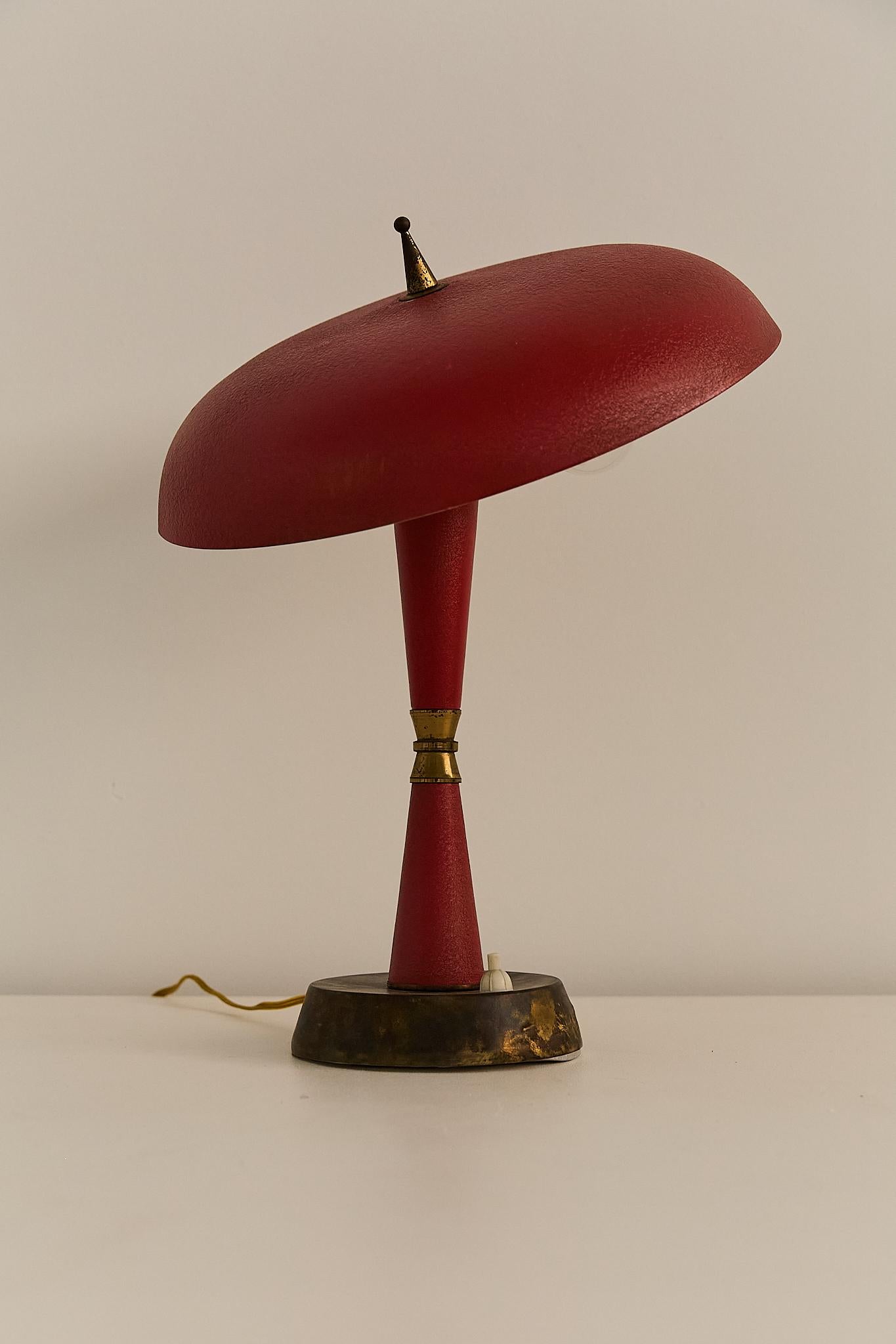A sculptural table lamp from 1950s Italy, featuring a bold red conical stem and wide saucer-shaped shade. The textured metal finish contrasts elegantly with brass accents, while the angled shade and exposed bulbs evoke classic space age and
