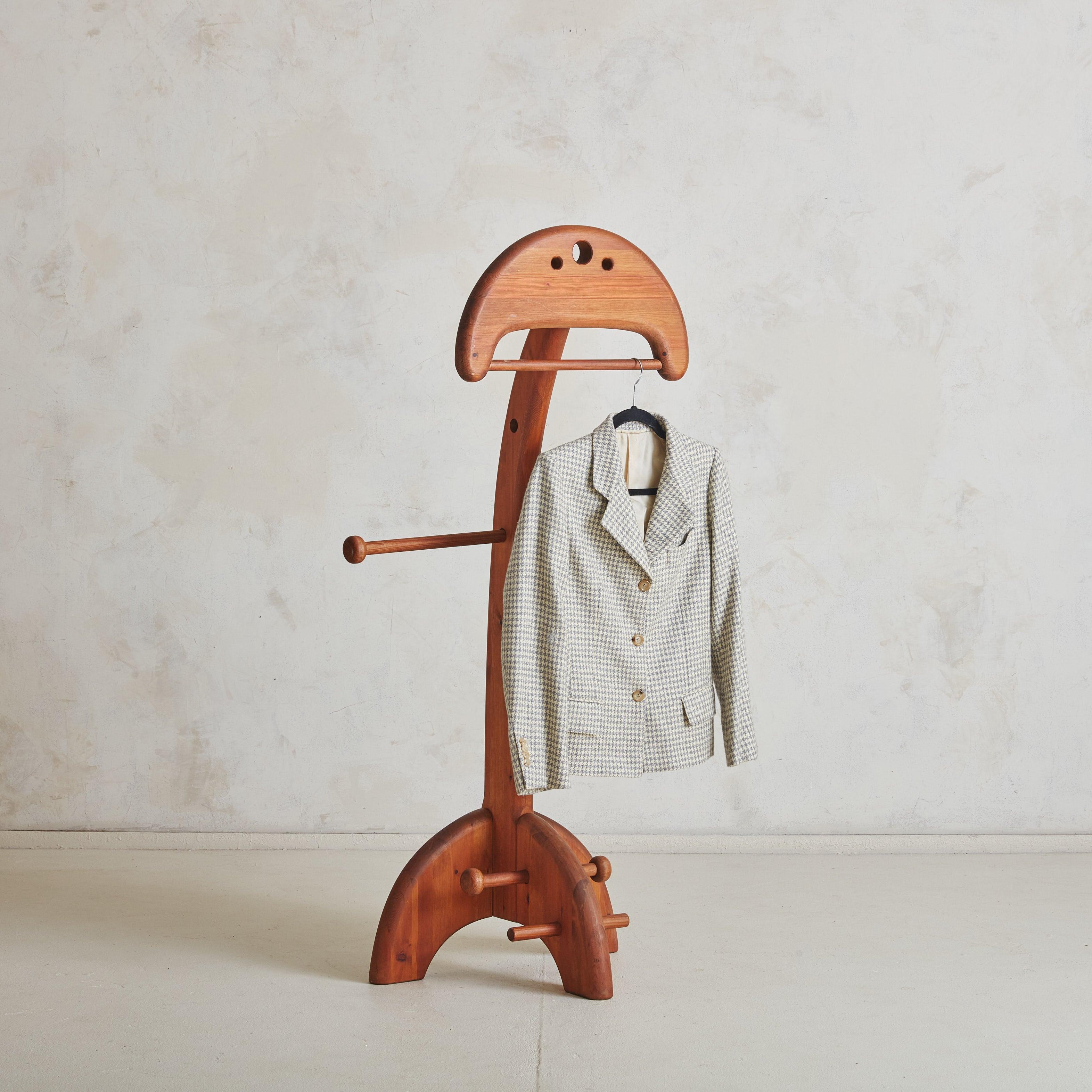 This sculptural teak valet from the 1960s captures the essence of mid-century modernism with its clean lines and organic warmth. The arched frame supports two hanging rods, one adjustable for larger garments, while the base offers a clever platform