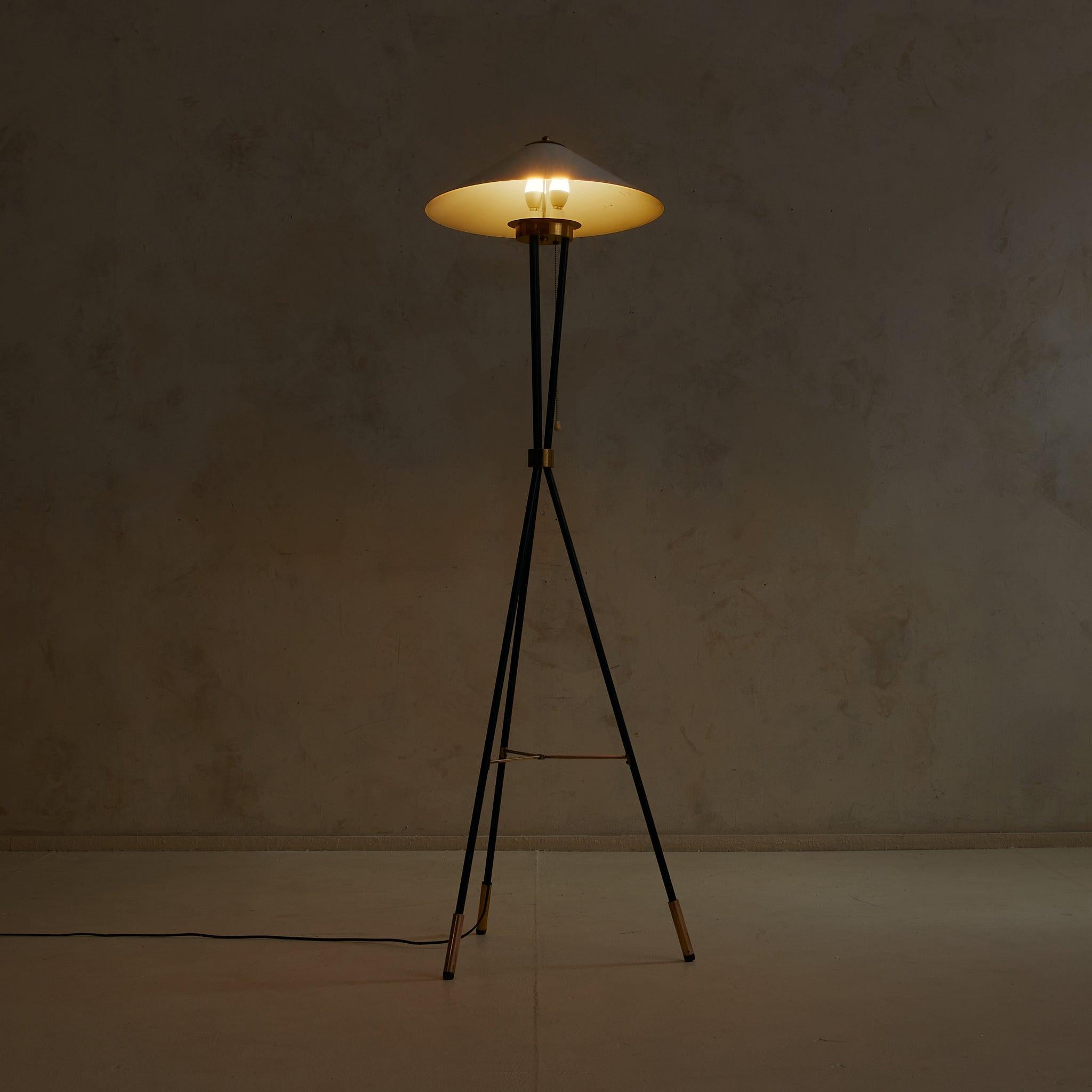 A 1950s Italian floor lamp with a sculptural tripod silhouette. The white-lacquered aluminum shade flares outward in a broad cone, resting above a brass collar and three slender black enameled metal legs. Polished brass accents punctuate the