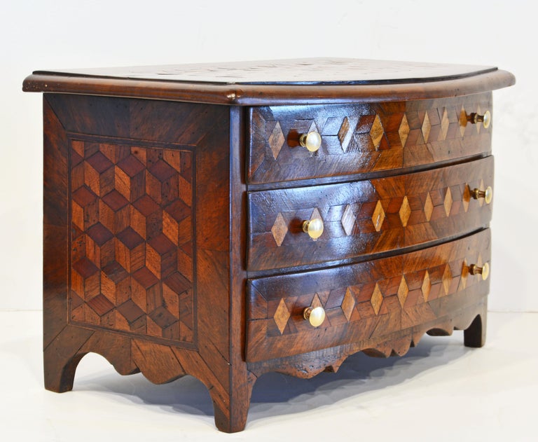 Dating to the early 19th century this gem of a miniature bow front commode or jewelry box features intriguing tumbling block parquetry on the top, the drawer front and the two sides. The commode rests on four bracket feet joined by elegantly shaped