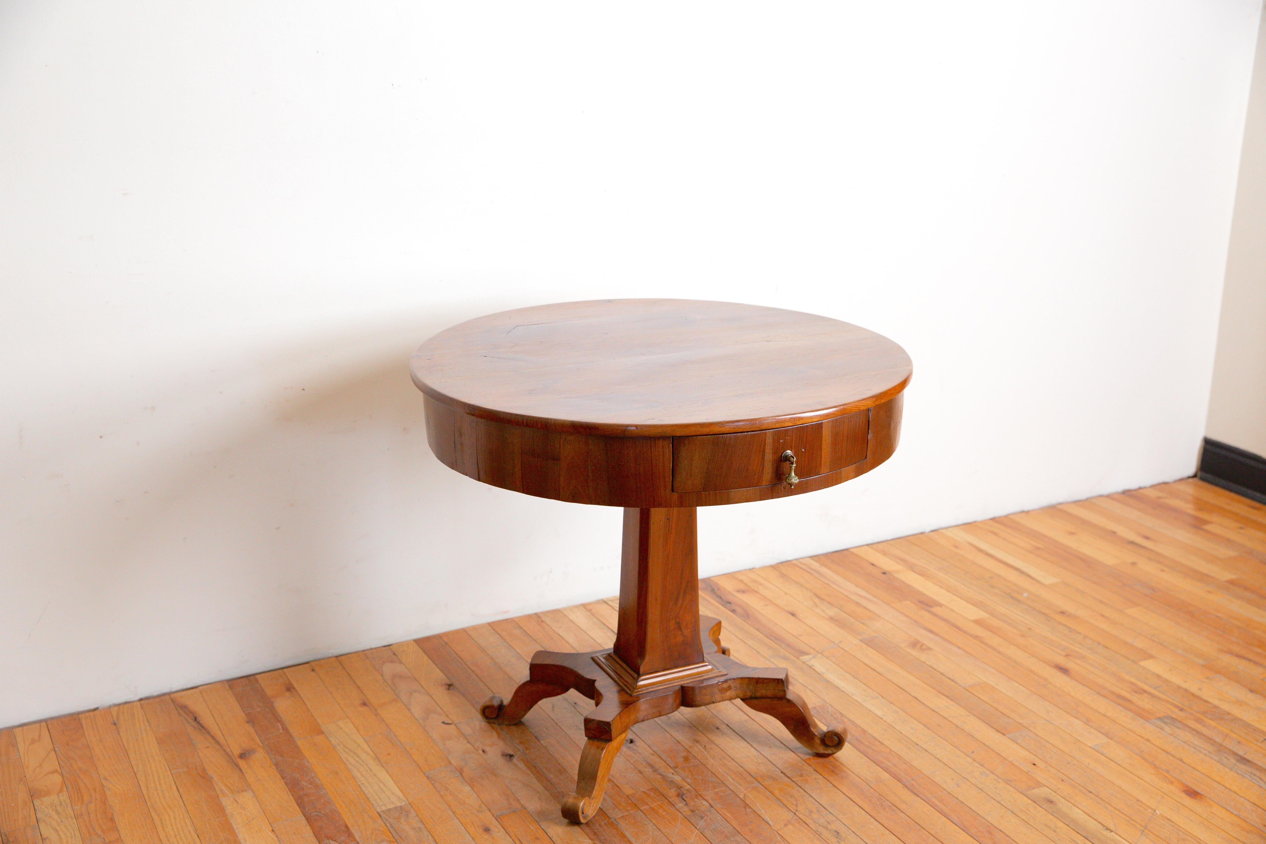 having a circular top above a conforming case housing one drawer raised on a slightly tapering 4-sided pedestal atop a shaped quadripartite base issuing four scrolled feet