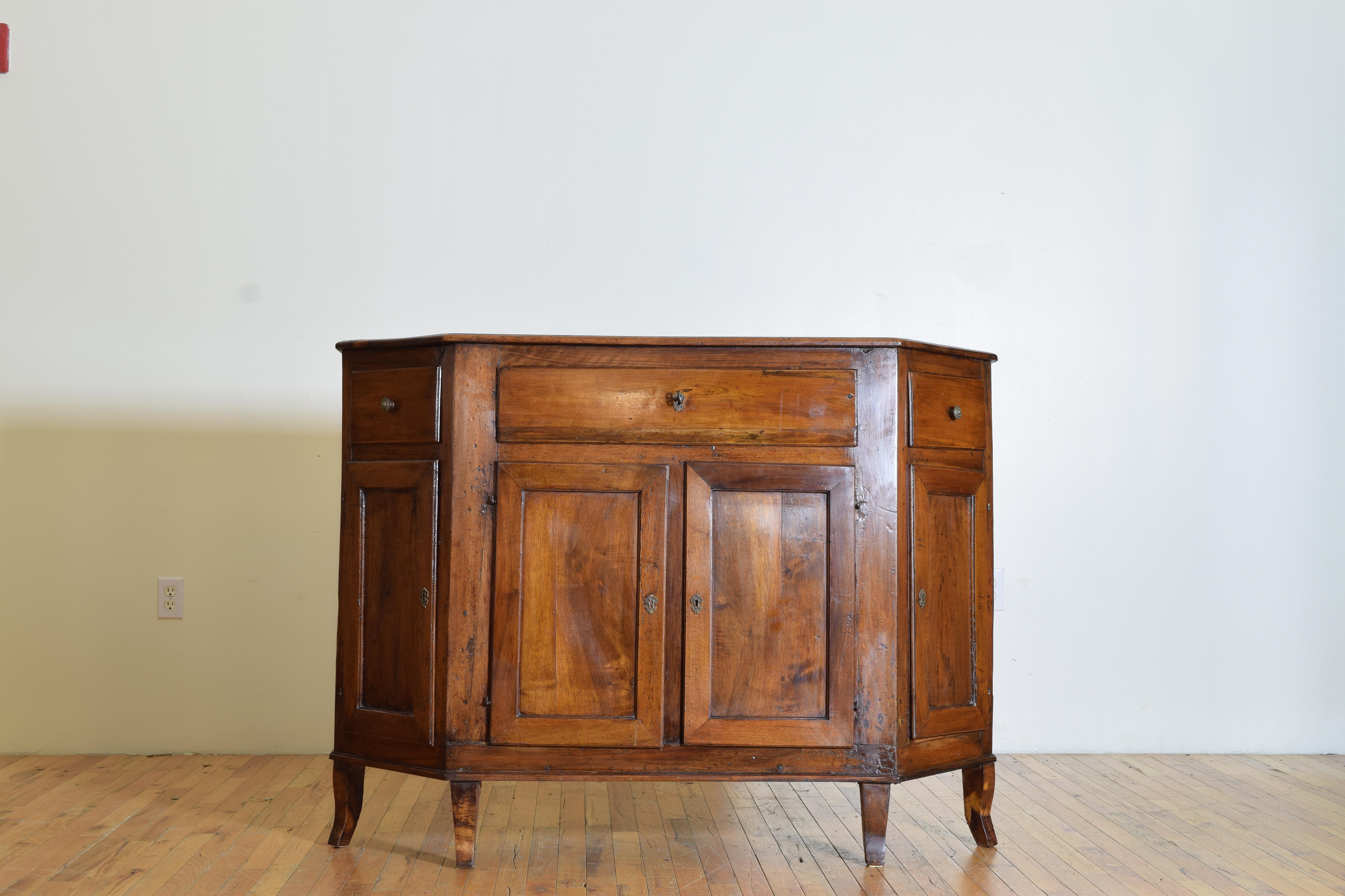 Having a shaped walnut top with canted “scantonata” profile and a molded edge above a conforming case housing one large drawer flanked by two smaller angled drawers, the lower part of the case with four doors opening to reveal shelving, raised on