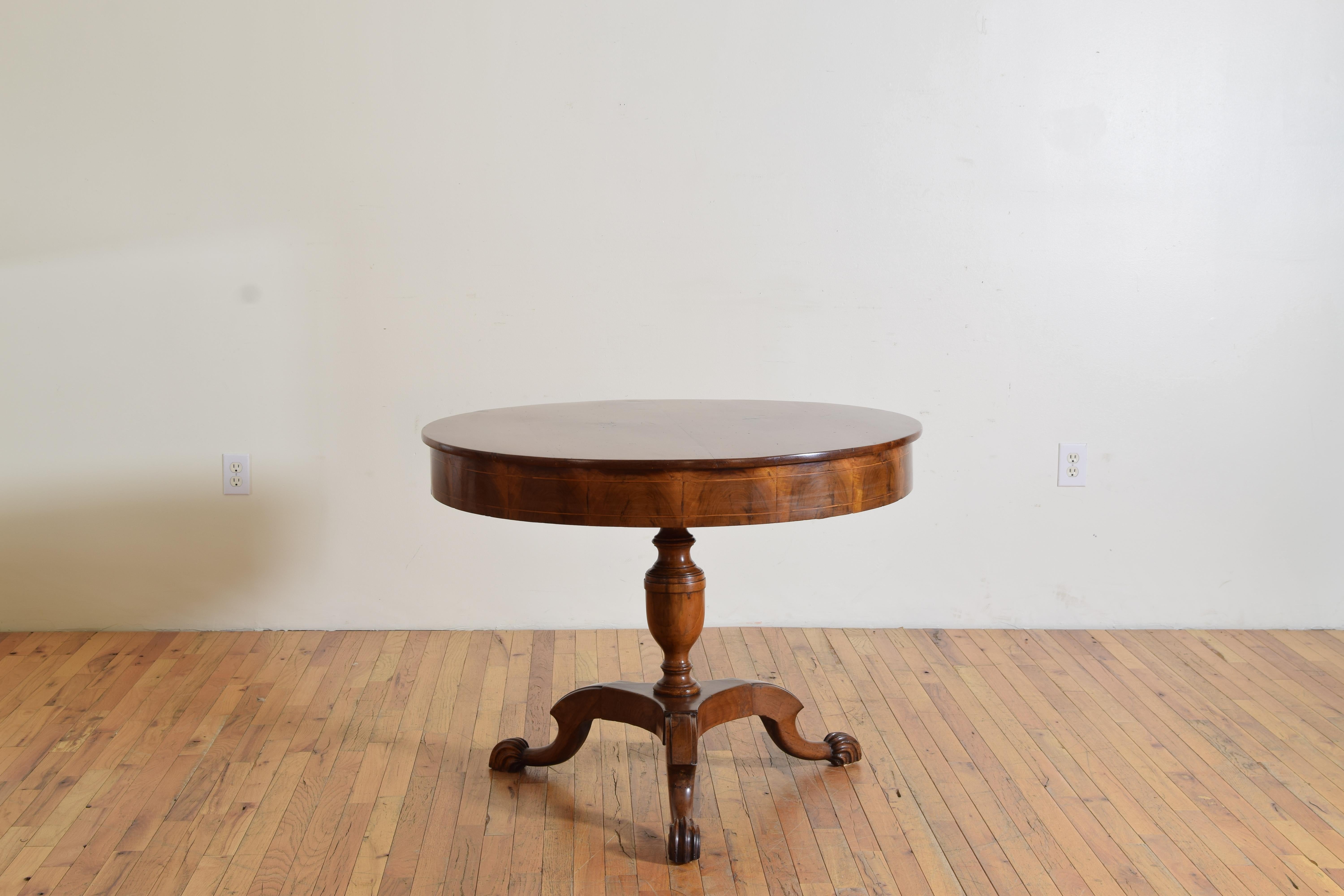 Having a round solid walnut top with an apron veneered in burl walnut with a double band of fruitwood inlay, raised on an urn-form turned standard atop a tripartite base issuing three shaped and expertly carved legs with shell carved feet