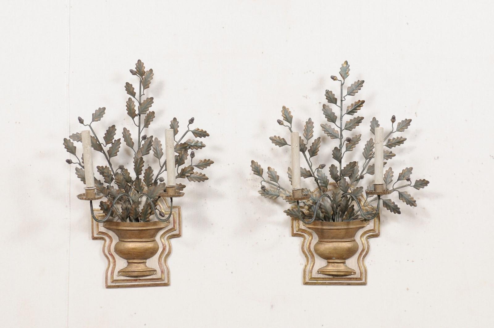 A pair of Italian two-light metal and carved wood candle sconces. This vintage pair of decorative wall sconces from Italy feature urn-shaped carved wood wall plaques as backplates, from which a metal arm swoops outward and in separation presents a