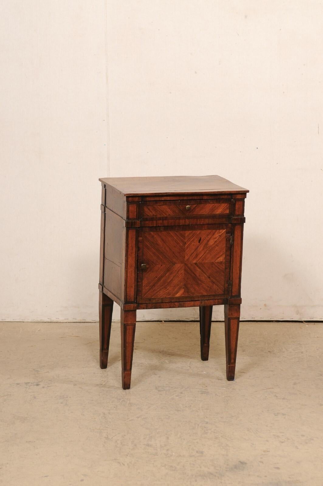 An Italian smaller sized veneered wood raised cabinet from the early 19th century. This antique end table from Italy has a case which houses a slender drawer at top, with a single door below (with open storage within). The chest has a beautifully