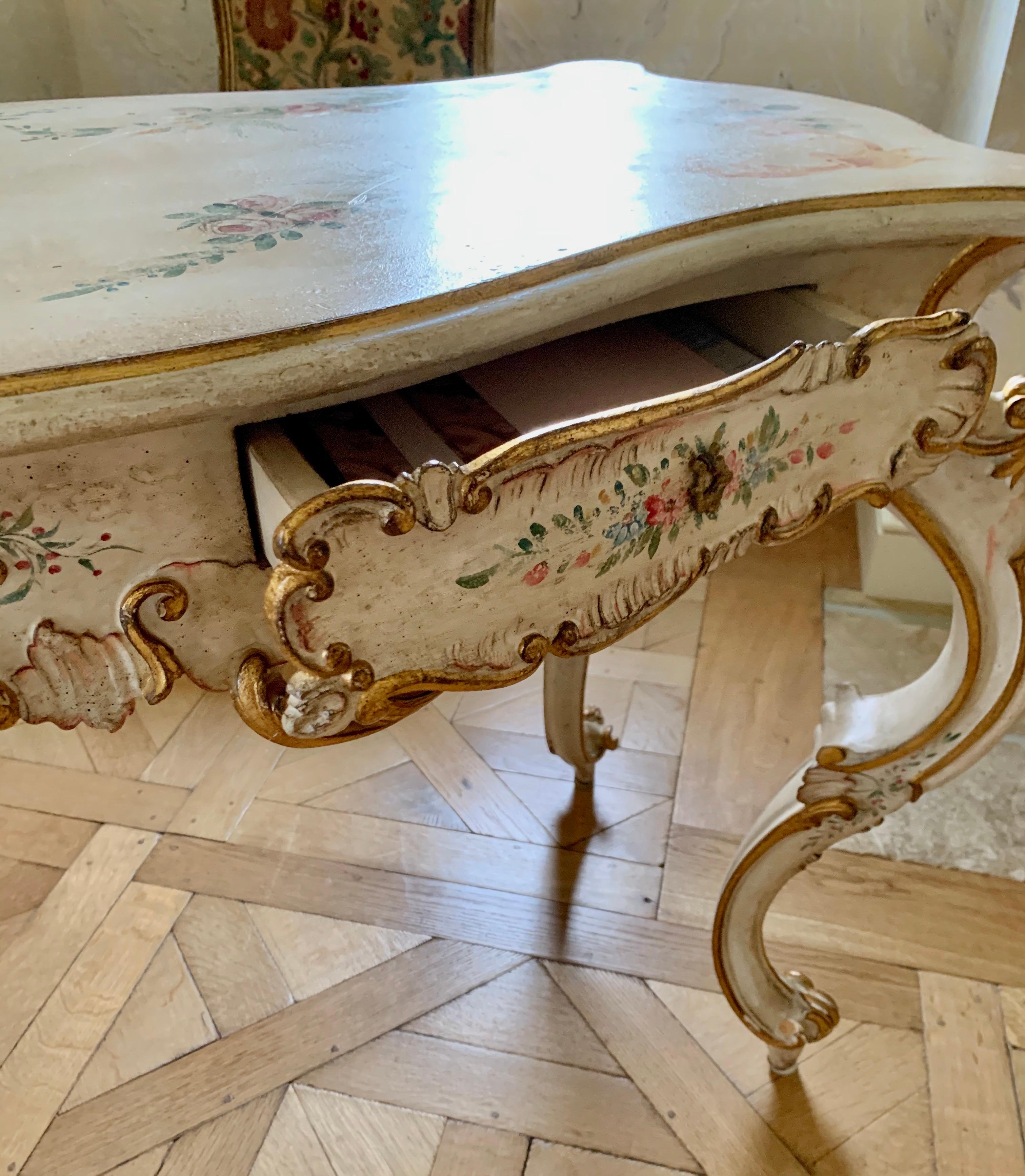 Exquisite hand painted ladies Venetian writing desk or dressing table with gold gilt carved scrollwork and hand painted floral motifs on all four sides. Desk has two drawers and one cabinet.