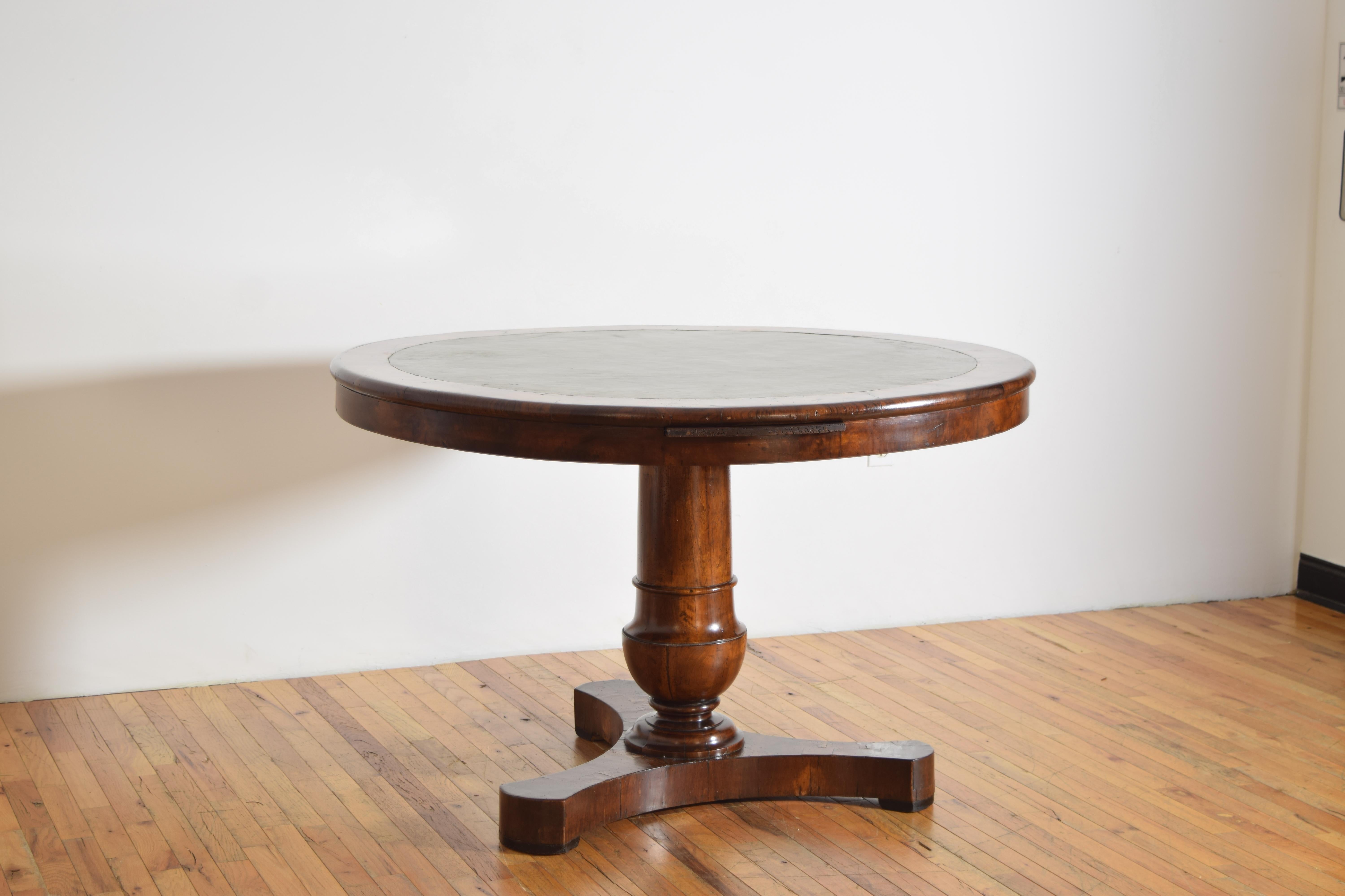 having a round top with rounded edge the center covered in leather, the conforming apron with a sliding writing surface also covered in leather, raised on a turned urn-form standard atop a tripartite base with shaped ends, raised on very small