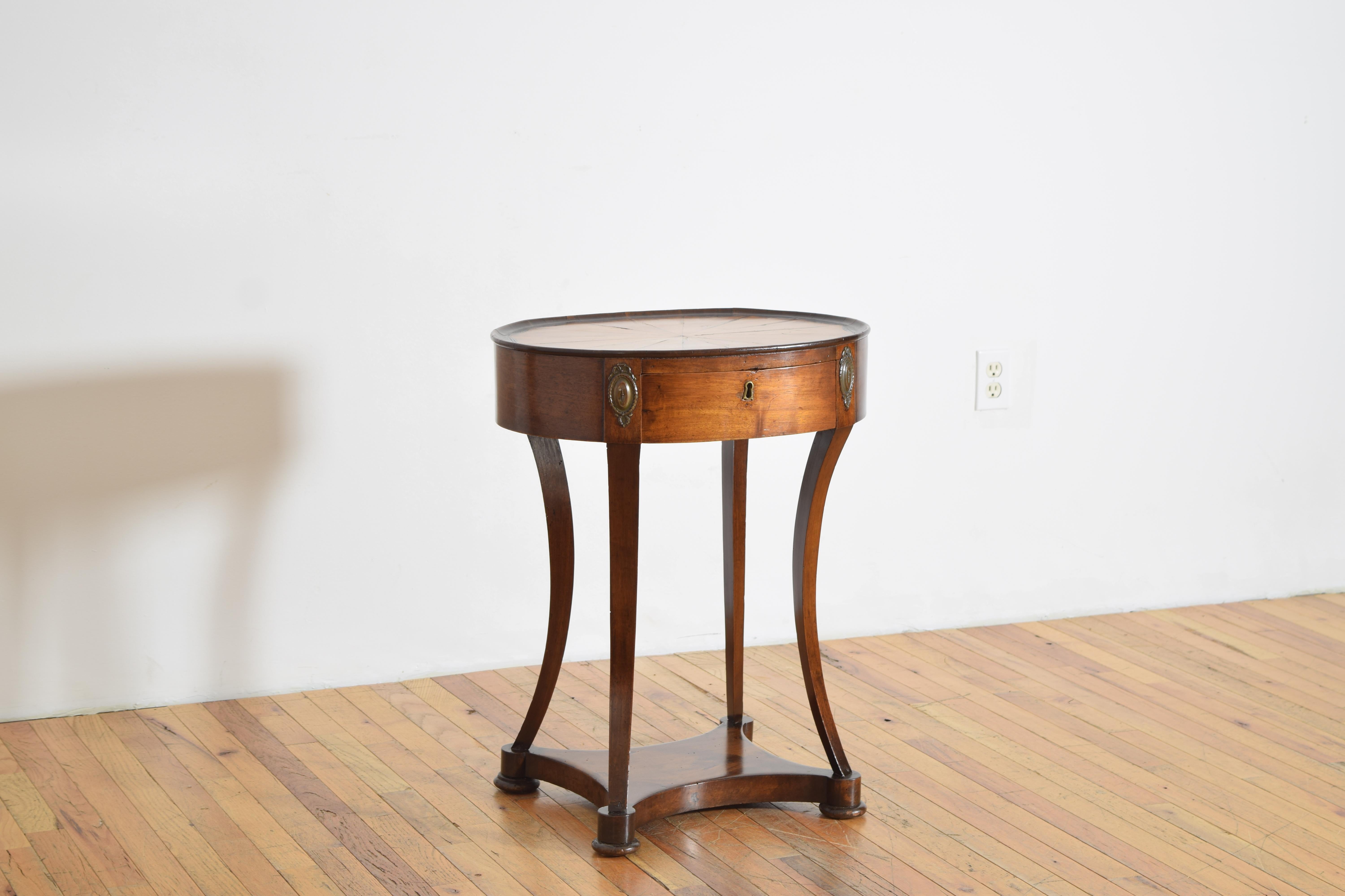 having an oval top with a radial ashwood and ebonized veneered surface and a slightly raised gallery, the case housing one drawer with a segmented compartments, the corners with patinated brass medallions, raised on saber shaped legs joining a lower