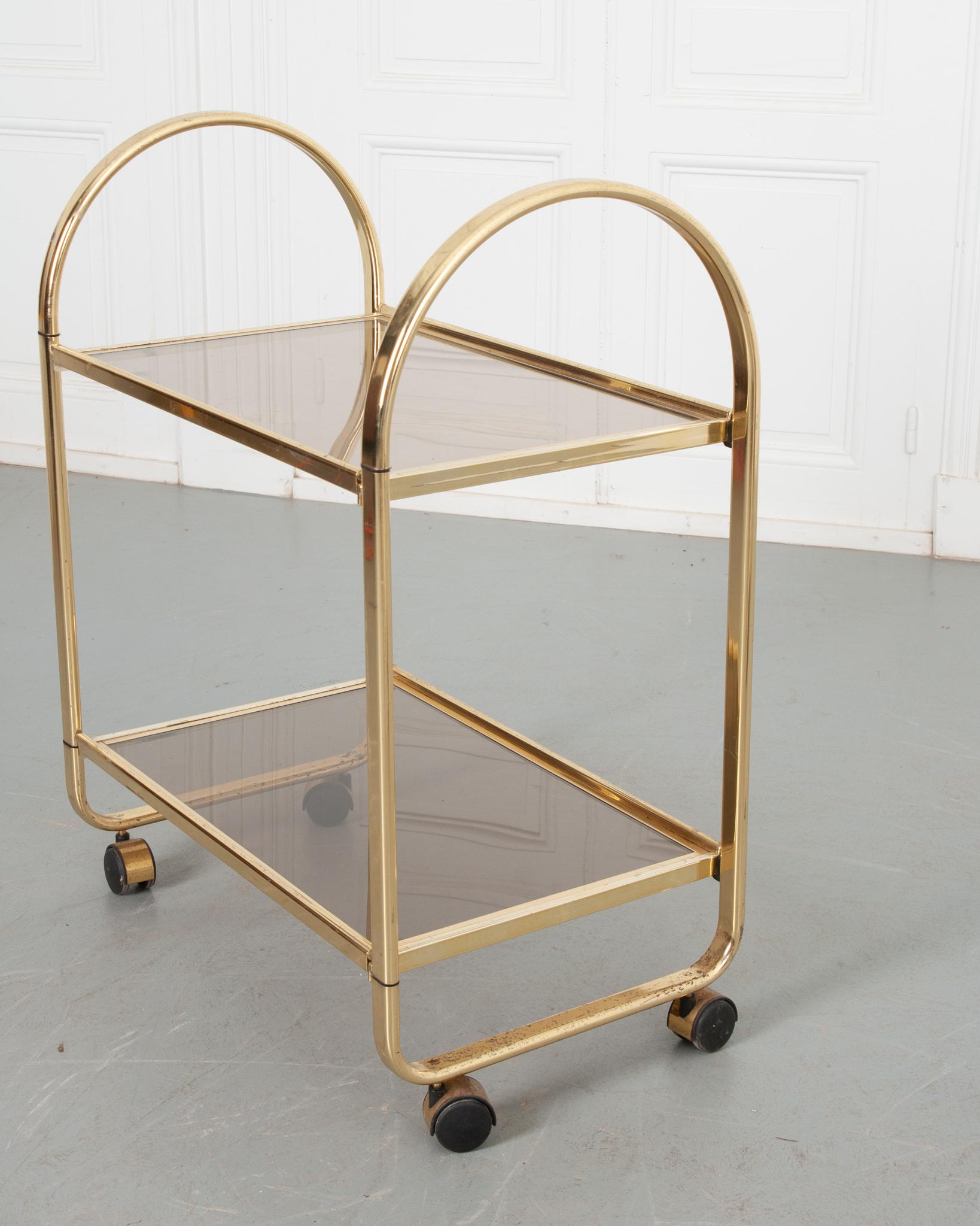 A charming petite bar cart crafted in Italy. The sleek metal frame retains a great luster that compliments the design. Two tinted glass shelves look wonderful from the side profile, are inset into the metal for a seamless look. Elevated on