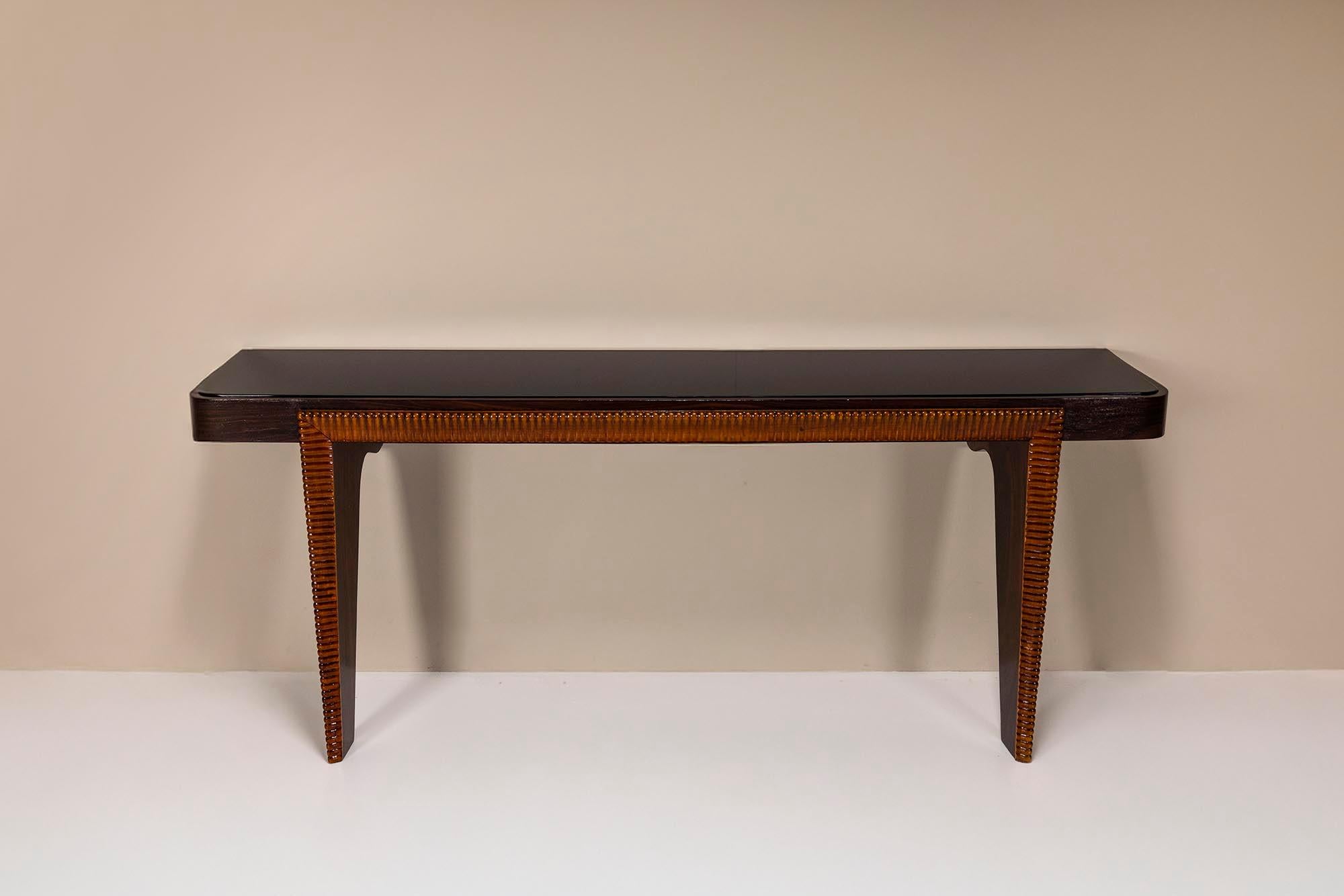 The console features a smoked black glass top, gently curved at the corners, which rests on a wooden structure. The dark finish of the wood contrasts beautifully with the warm tones of the finely carved decorative edge, running continuously along