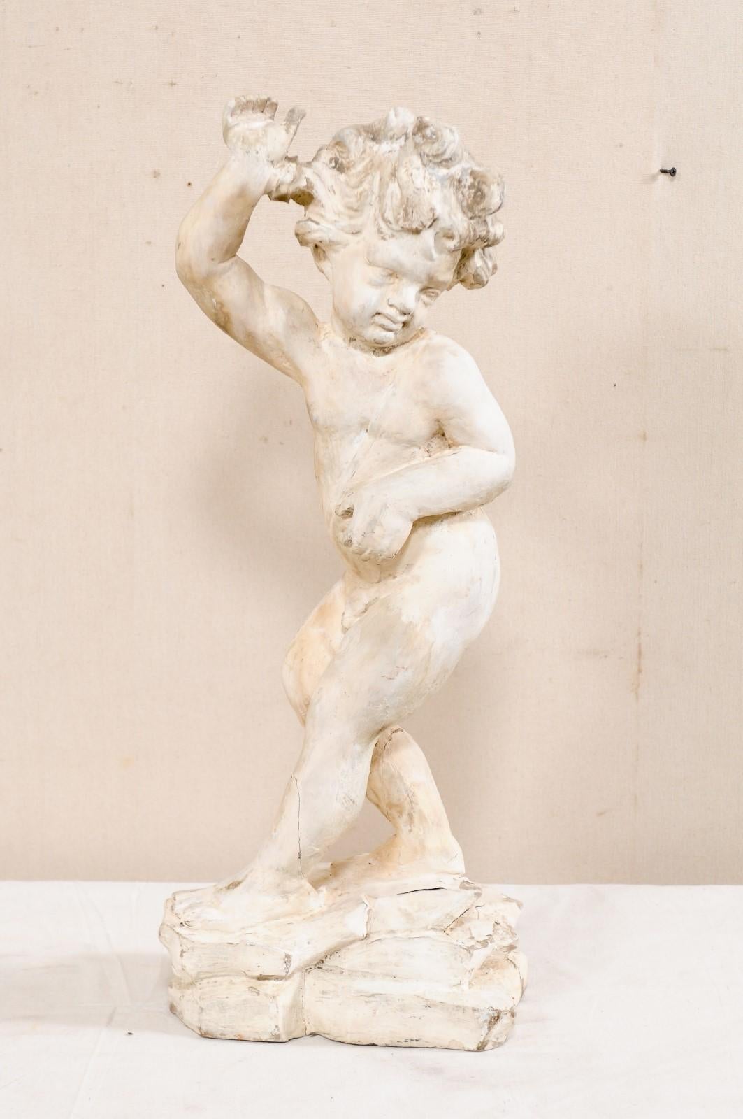 An Italian hand carved wooden putto figure. This vintage figure from Italy has been hand carved of wood and depicts a nude male child, standing in a twisted position as if turning or looking down-wards behind him, with his right arm and hand raised