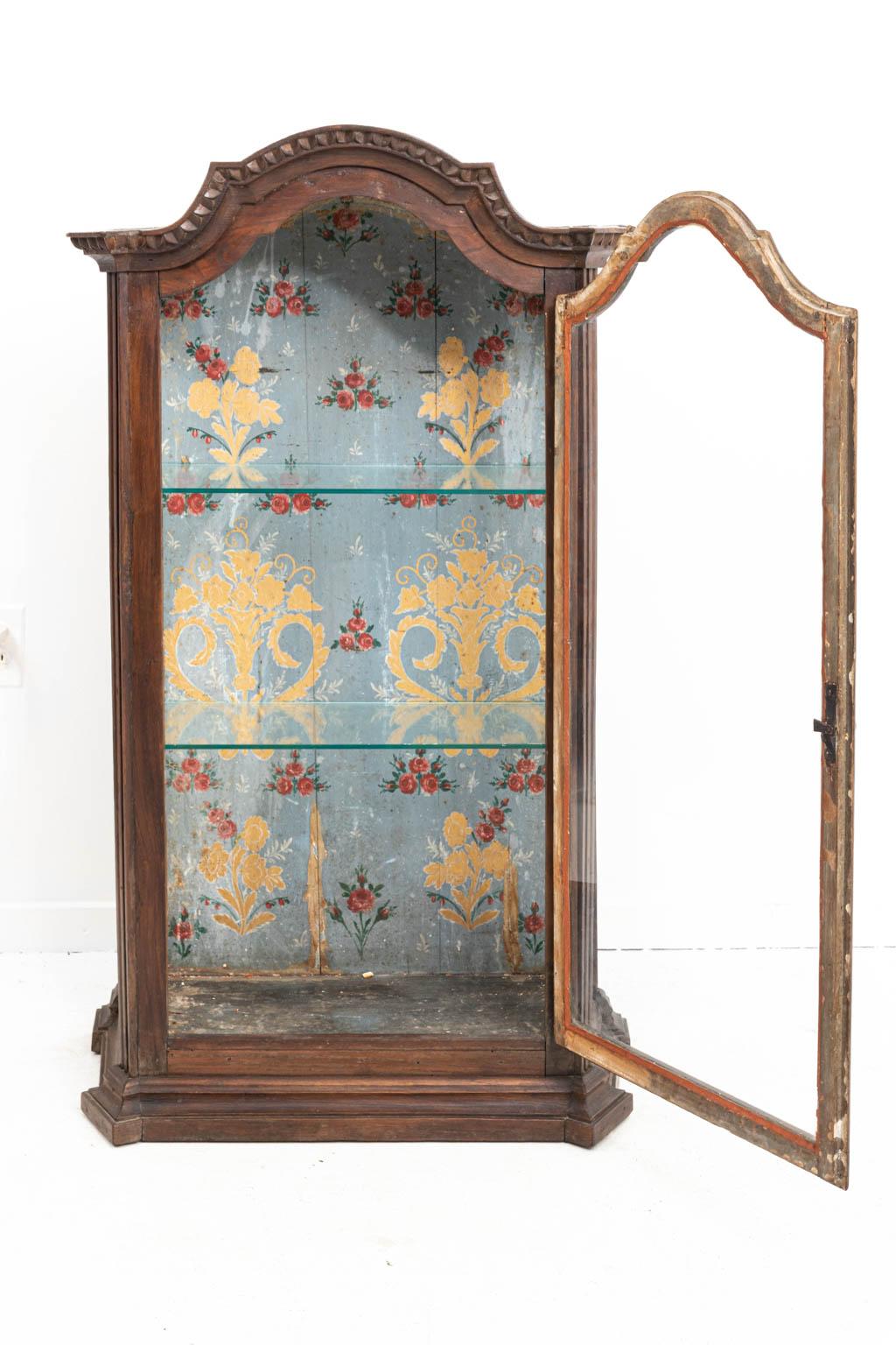 Italian vitrine wall cabinet in walnut with paint decorated interior of flowers and glass shelves, circa early 19th century. The piece also features a glass front. Please note of wear consistent with age including paint loss and finish loss to the