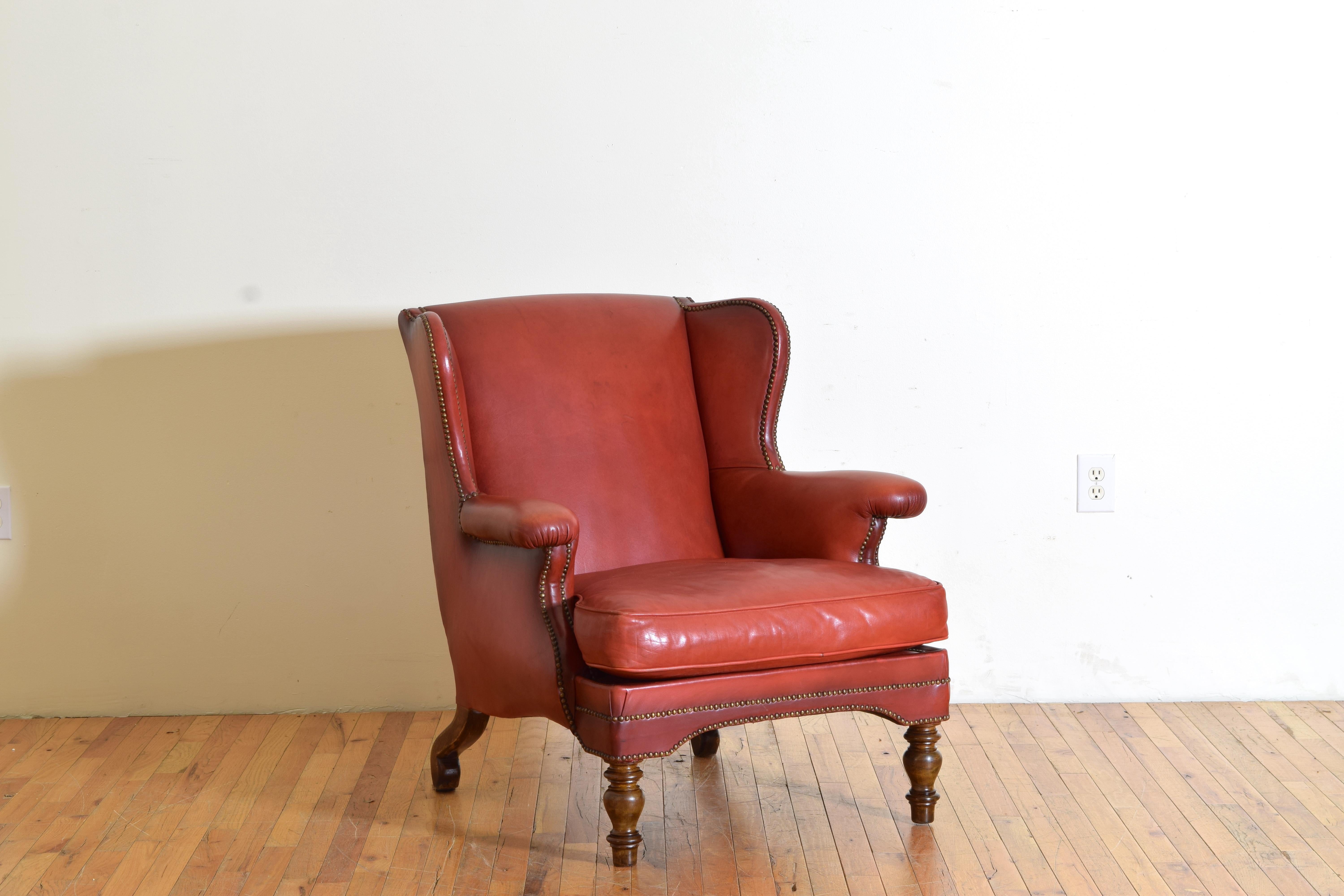 This deep set leather chair is completely covered in a faded red leather trimmed in patinated brass nailheads, the closed arms jutting forward slightly, with a loose cushion, raised on turned front legs and wonderfully shaped extended back legs