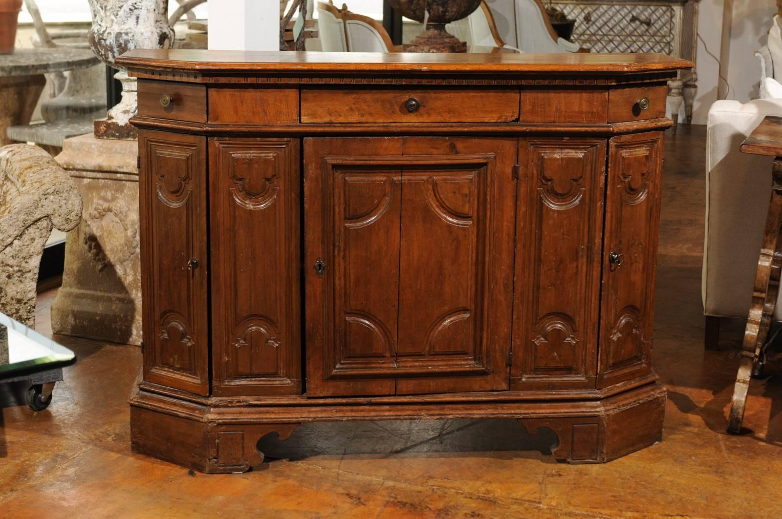 An Italian 19th century walnut narrow credenza from Siena with canted corners, geometrical motifs, three drawers and three doors. Born in Tuscany, one of the most enchanting regions of Italy, this walnut credenza features a nicely aged shaped top