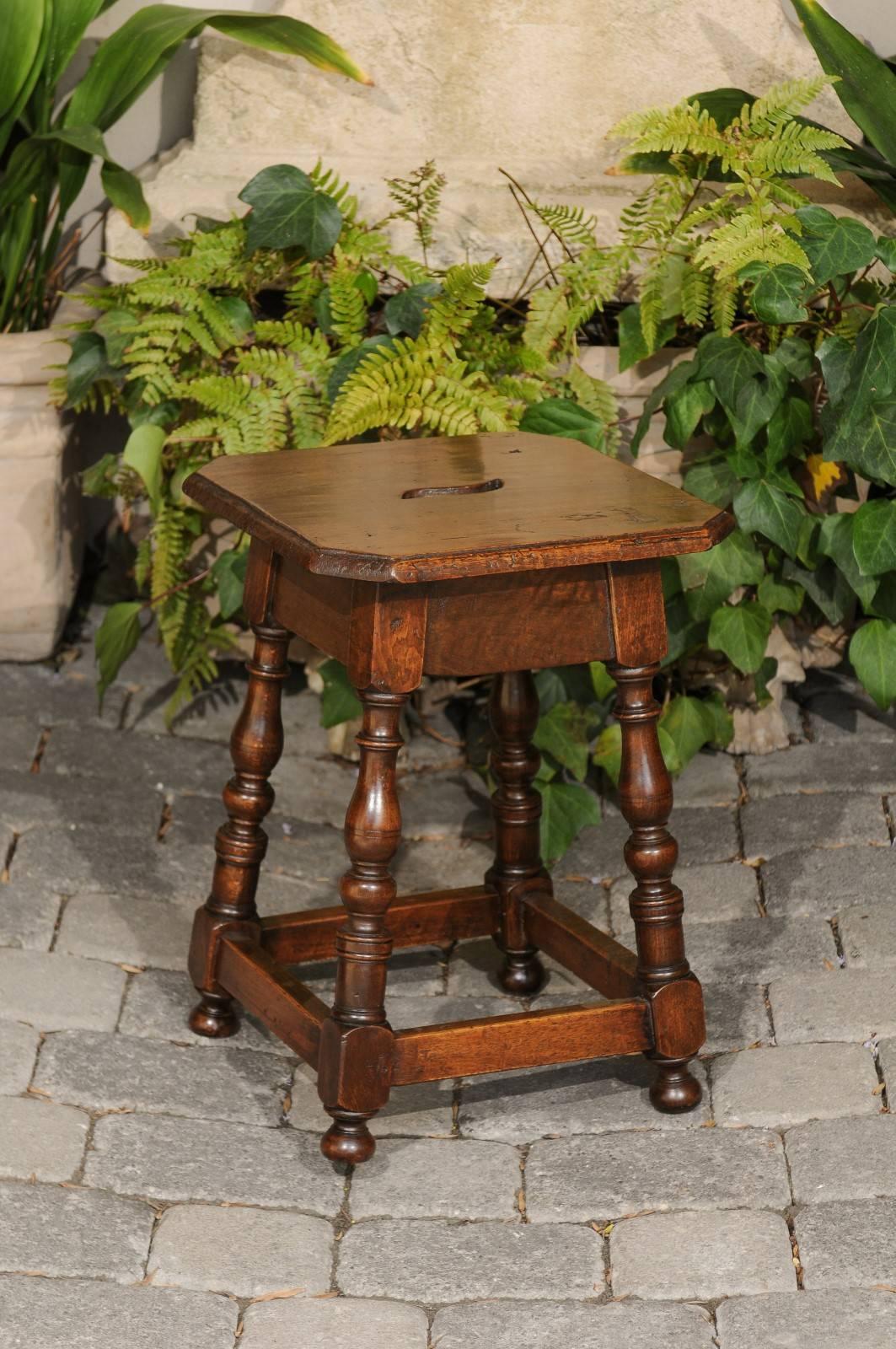 An Italian walnut stool with turned legs and side stretcher from the early 19th century. Born in Italy during the 1820s, this Italian stool features a square wooden seat with canted corners and pierced handle, resting on a simple apron. Four