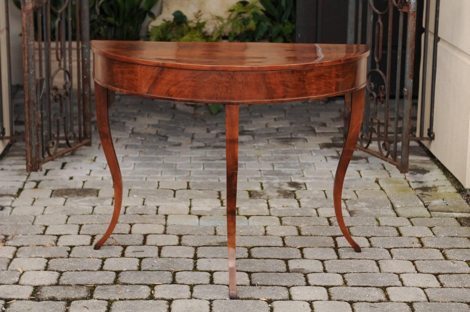 An Italian walnut demilune console table with tripod base from the first half of the 19th century. This Italian demilune features a semi-circular top, sitting above a bookmark veneered apron. The table is raised on three elegant curved legs, with
