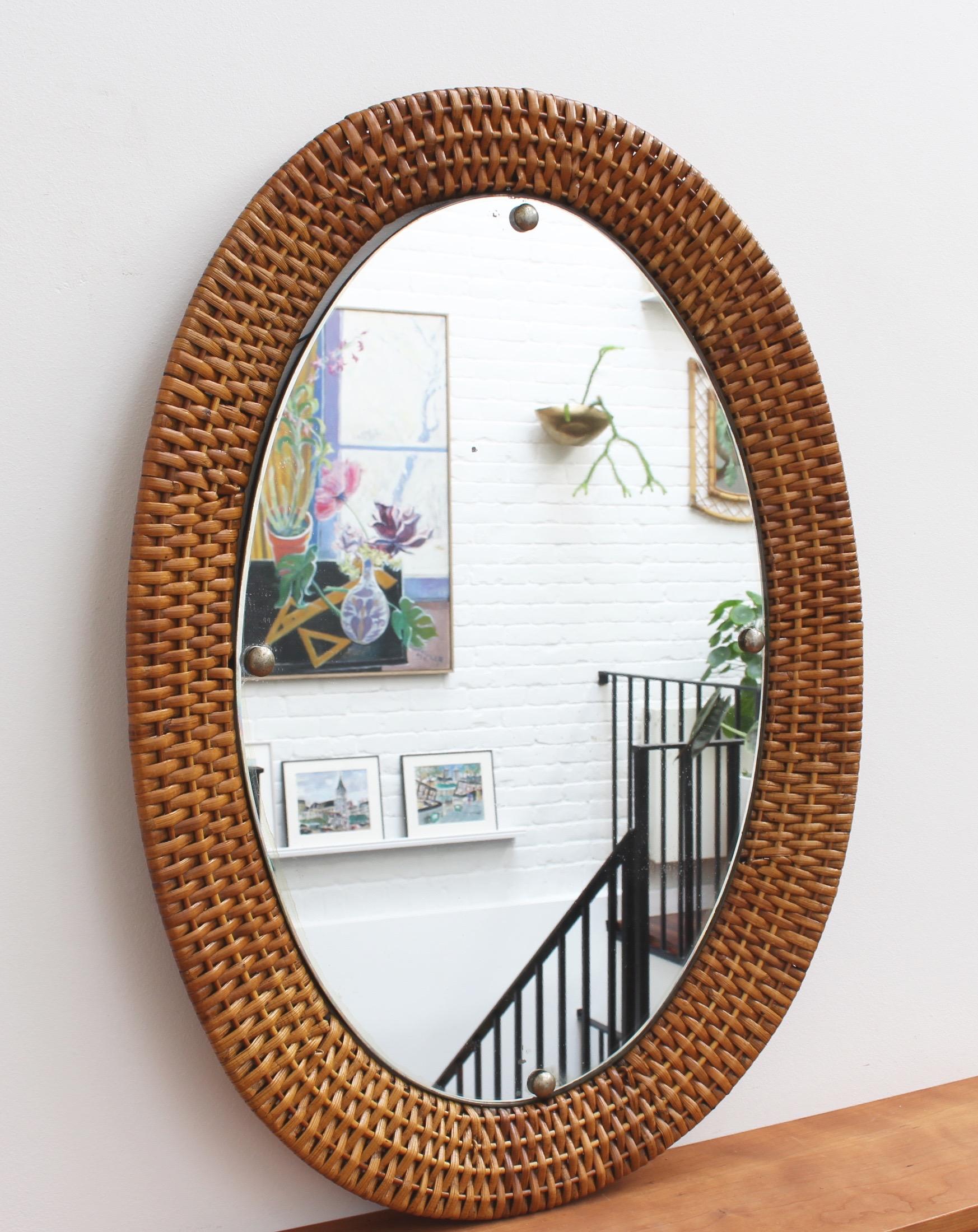 Italian wicker rattan wall mirror (circa 1960s). A distinctive vintage mirror which transports you immediately to another place and another time, perhaps the Italian Riviera or Miami's South Beach circa 1960. Its substantial glass is oval-shaped,